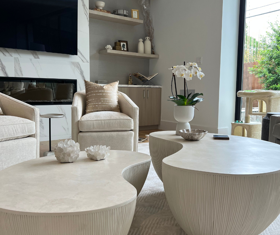 a bright living room with a pair of coffee tables, beige club chairs, built-ins, and a stone fireplace surround