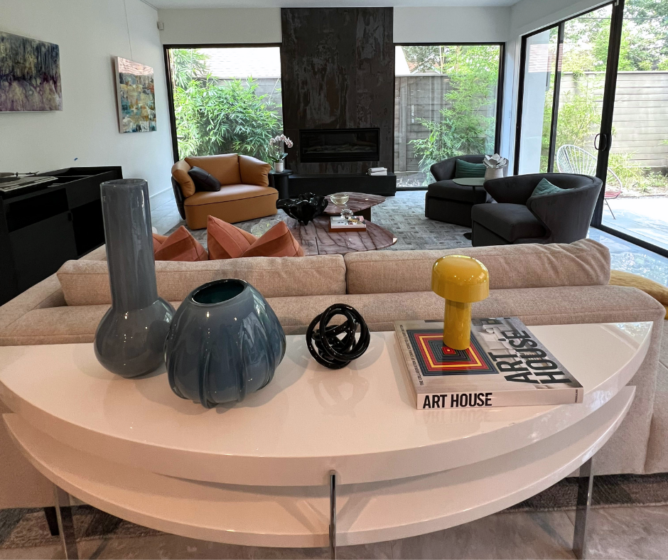 a living room with club chairs, a large slab fireplace, couch, and console table, vessels, books, and decor objects on top