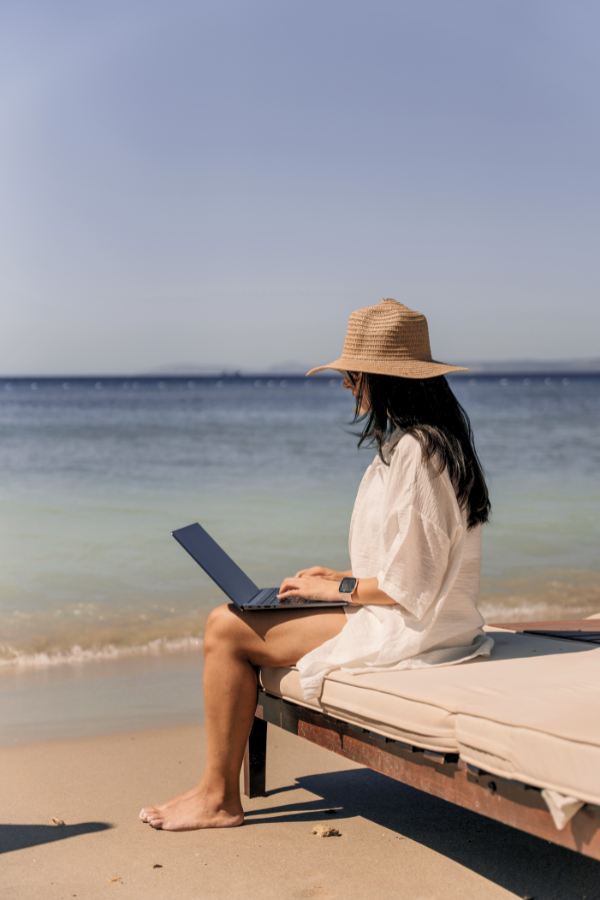 a woman wearing a hat working on her laptop on the beach