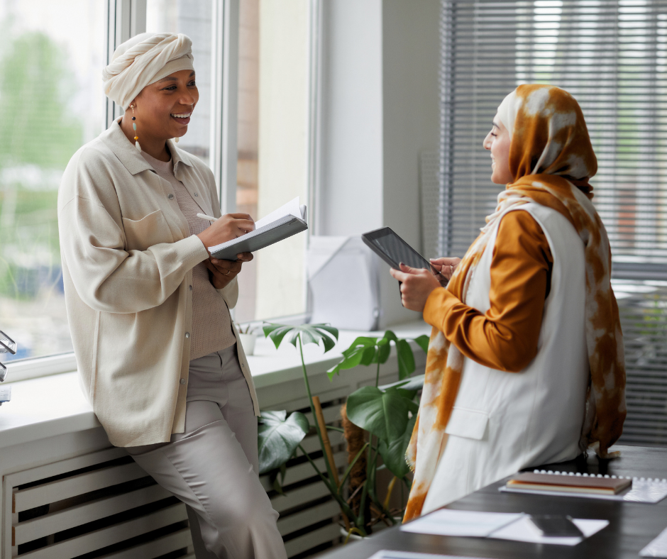 two women hold tablets and chat with each other in front of a window