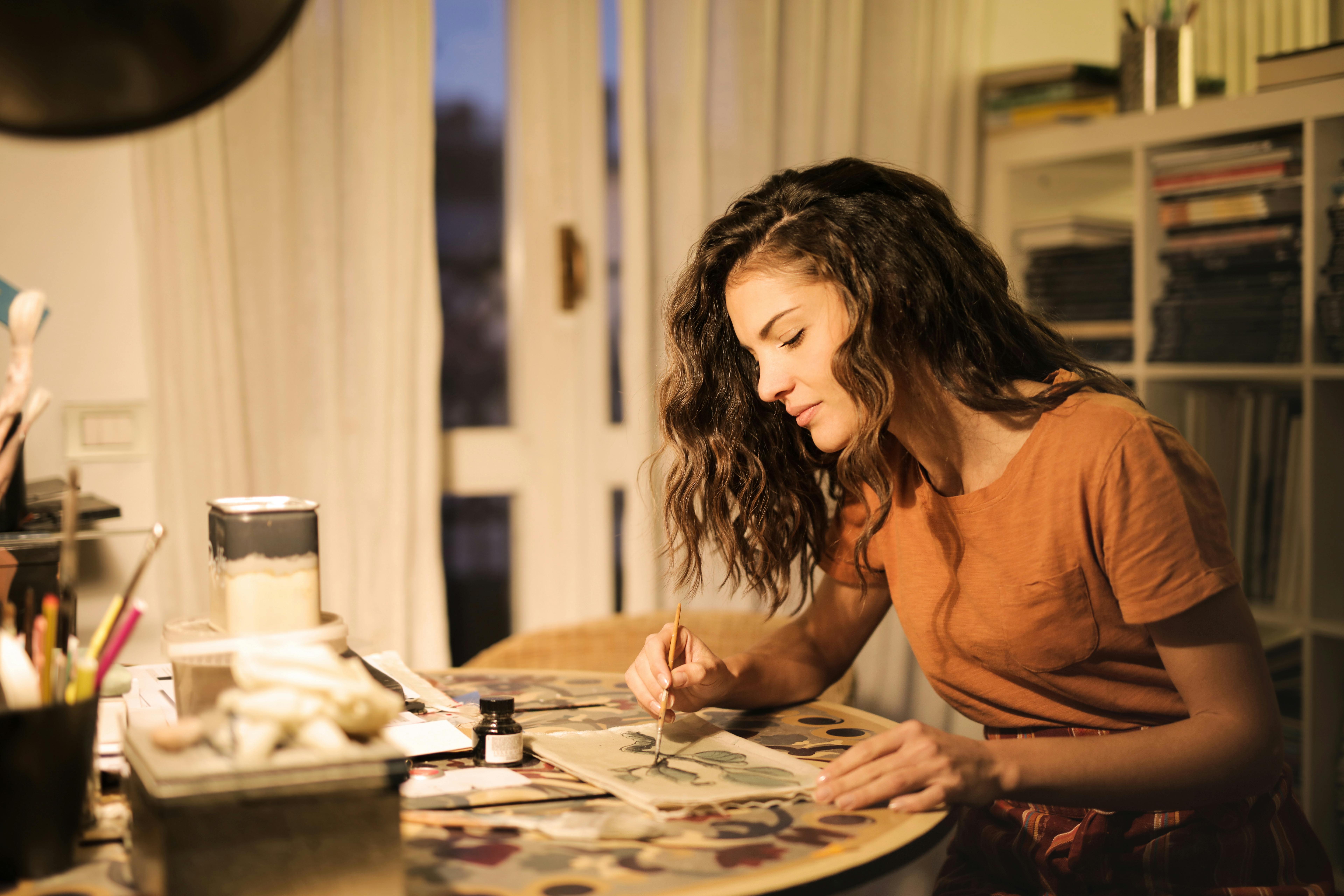 A woman painting at a table in a cozy indoor setting.