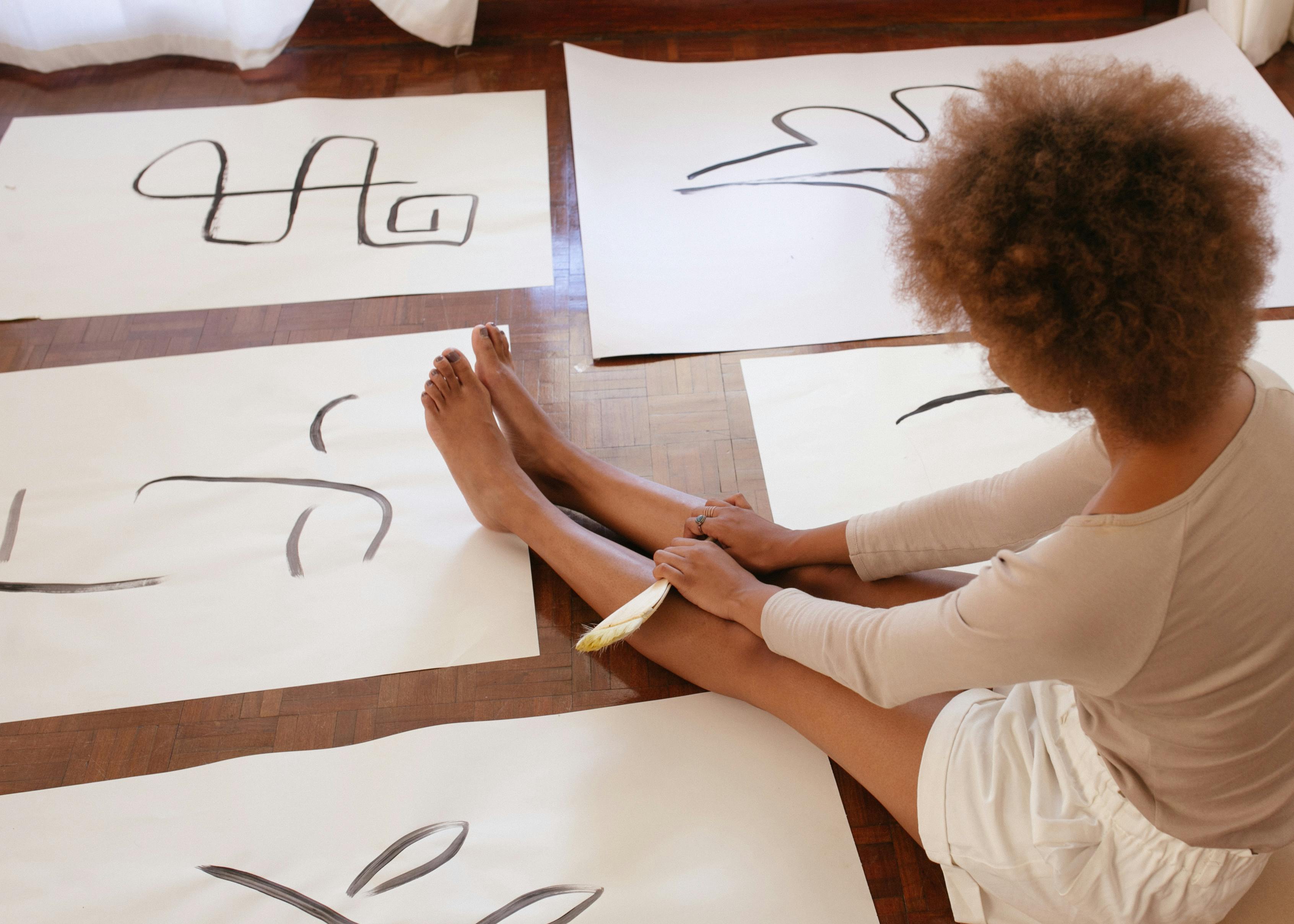 Person sitting on the floor surrounded by large sheets of paper with black brush strokes, using a feather brush.