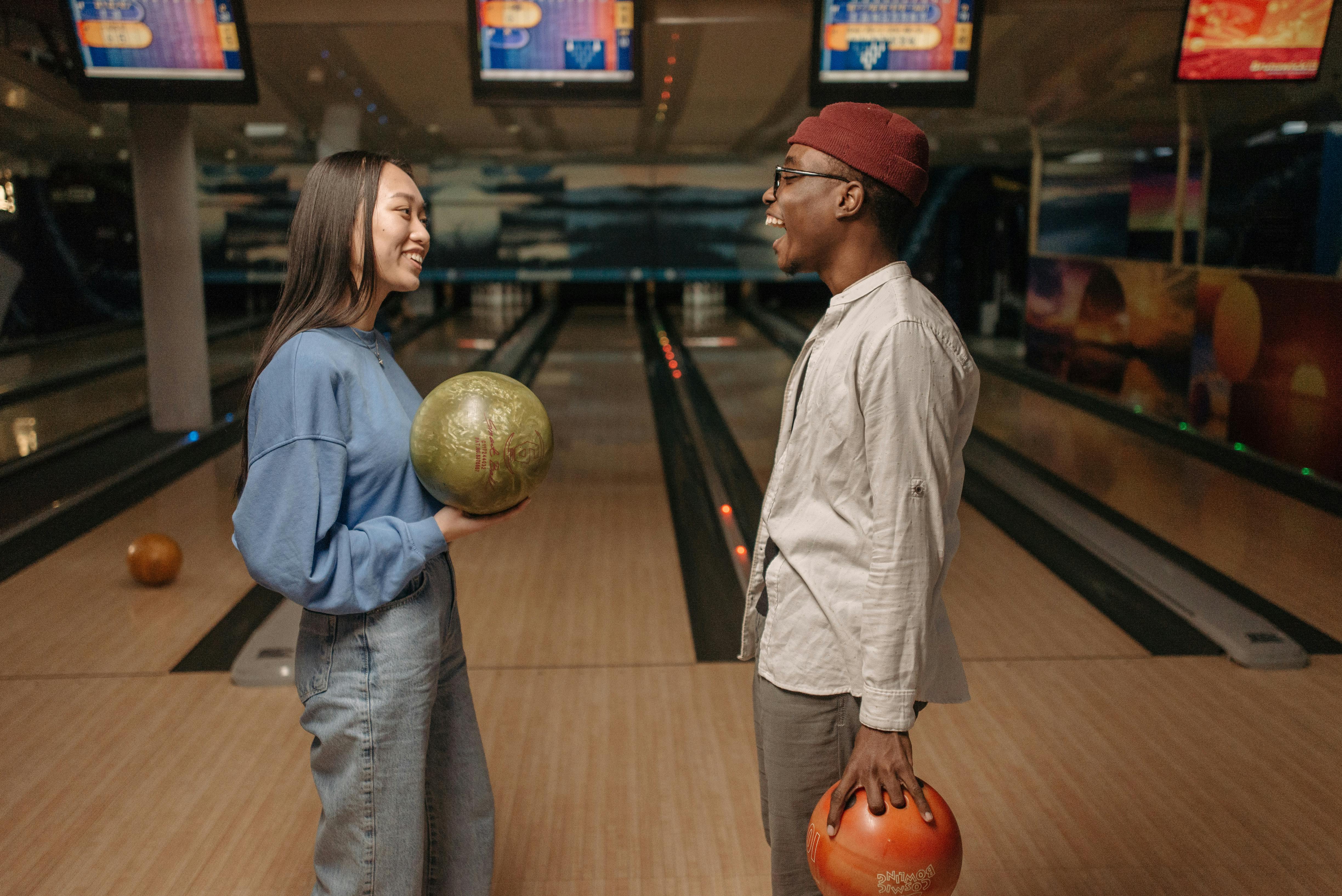 Two friends smiling and talking while holding bowling balls in a bowling alley.