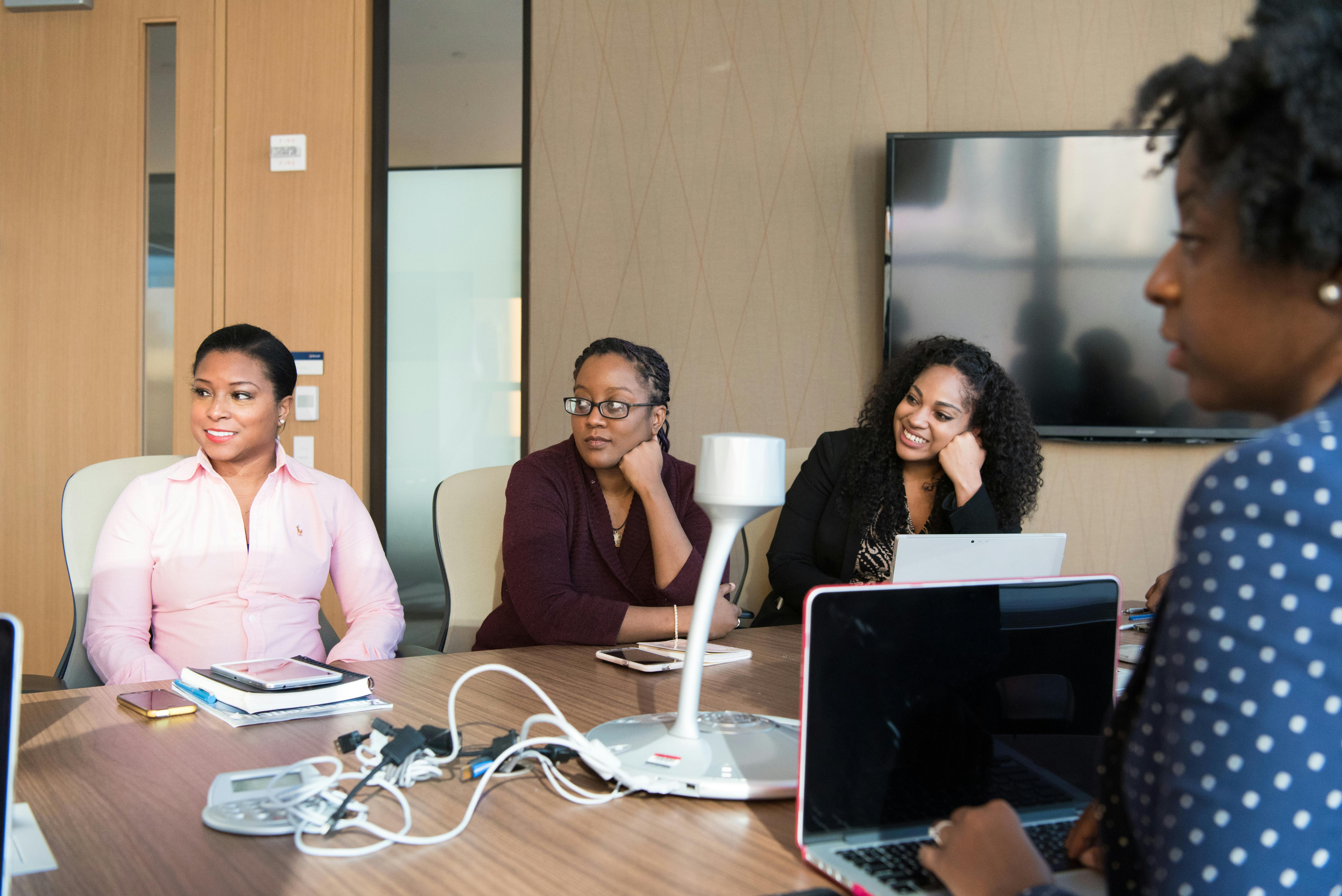 A group of diverse women engaged in a business meeting around a table with laptops and notebooks.