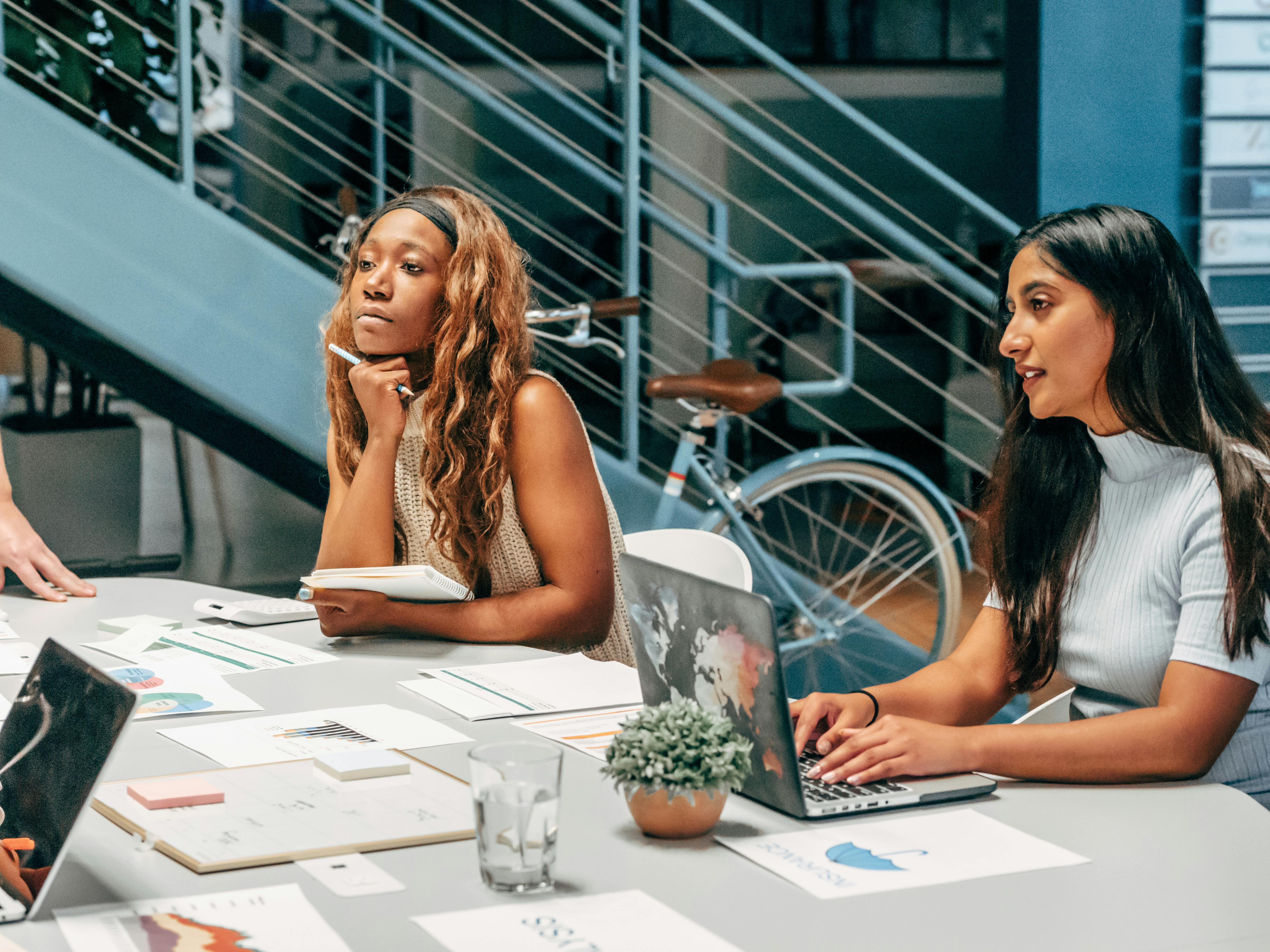 Two women engaged in a business meeting at a modern workspace with various documents and a laptop on the table.