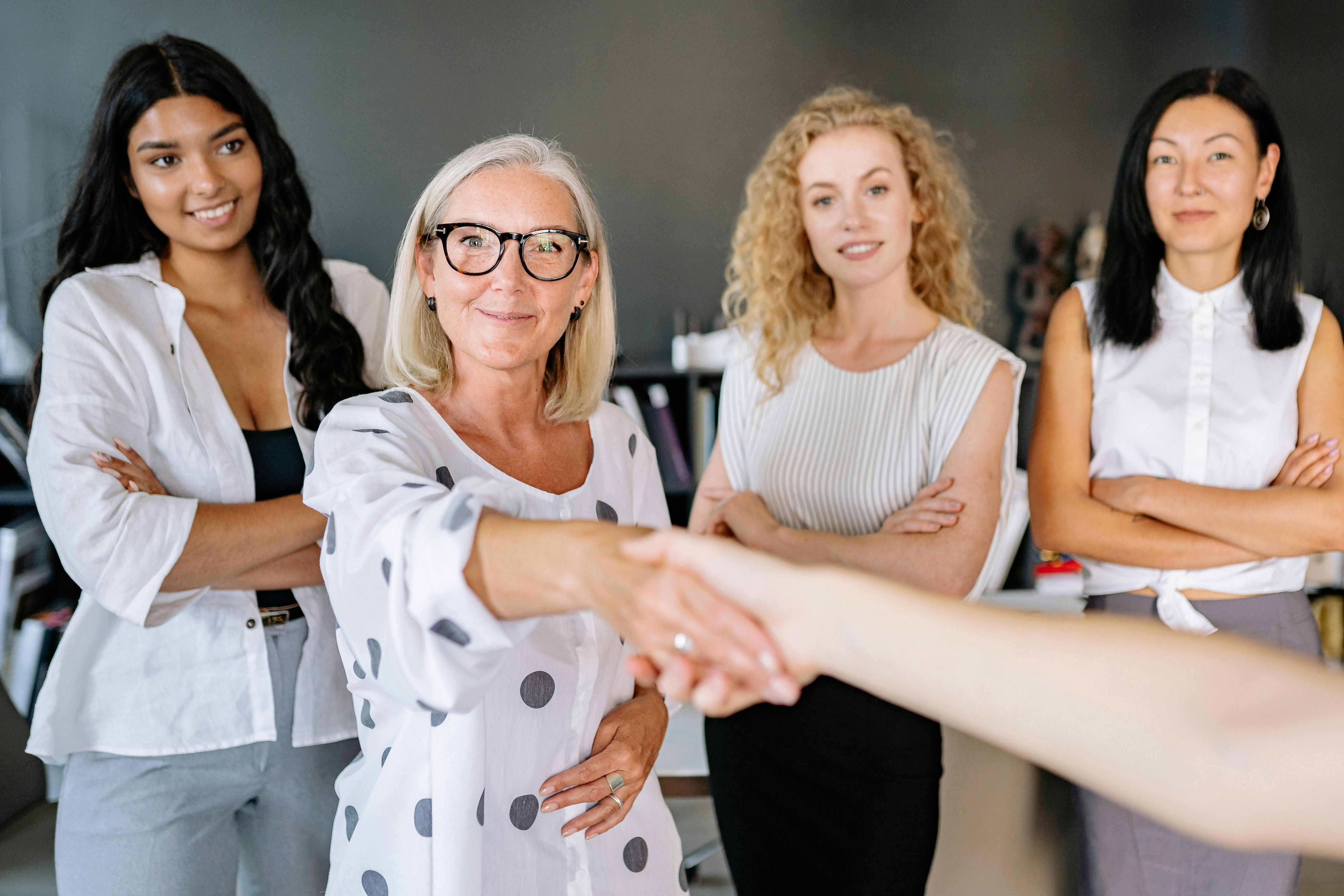 A group of four women in a modern office, with one woman shaking hands with another.