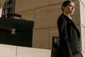 A confident businesswoman in a black suit, glasses, and a briefcase, standing against a stone background.