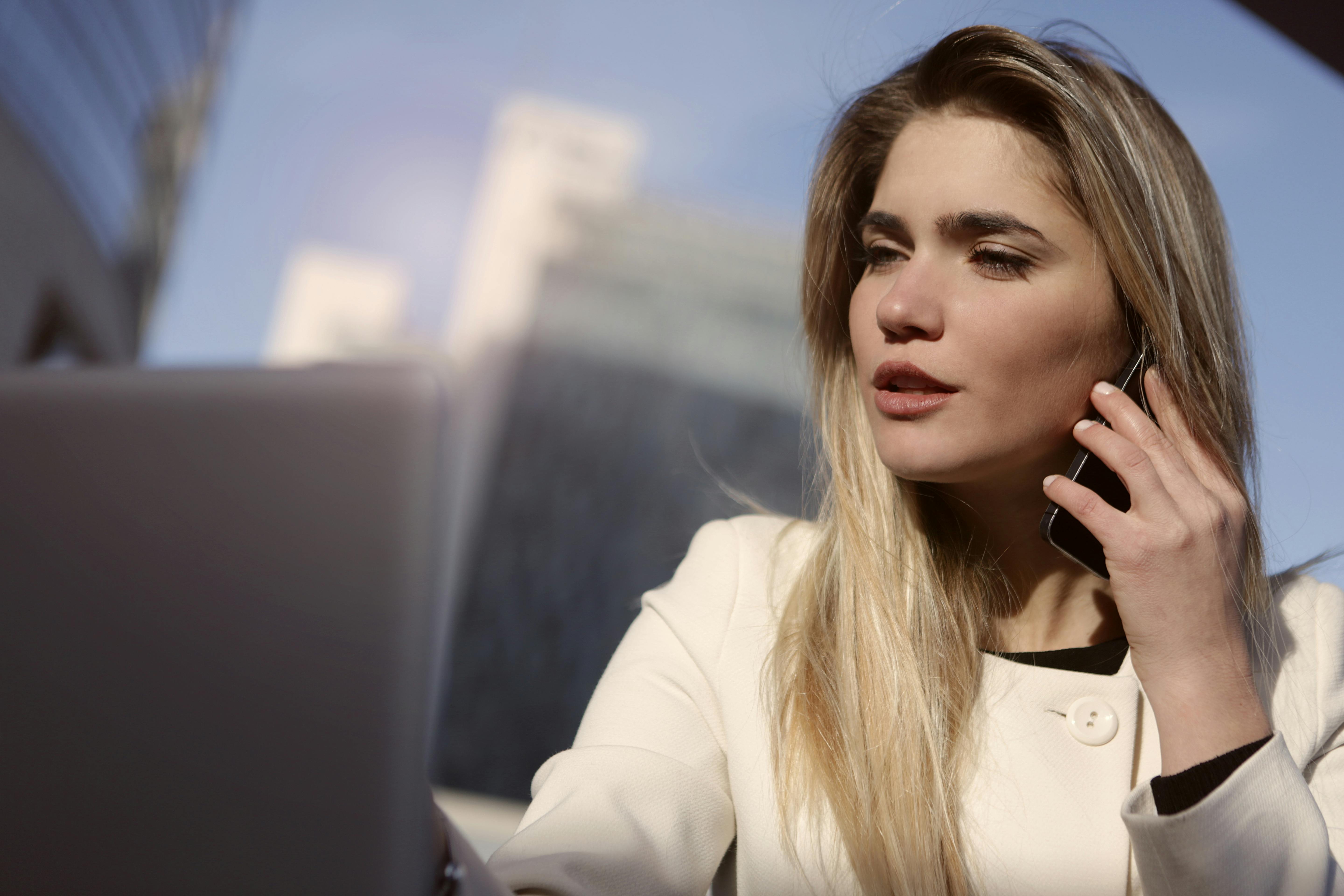 Woman in a white blazer talking on the phone while working on a laptop.