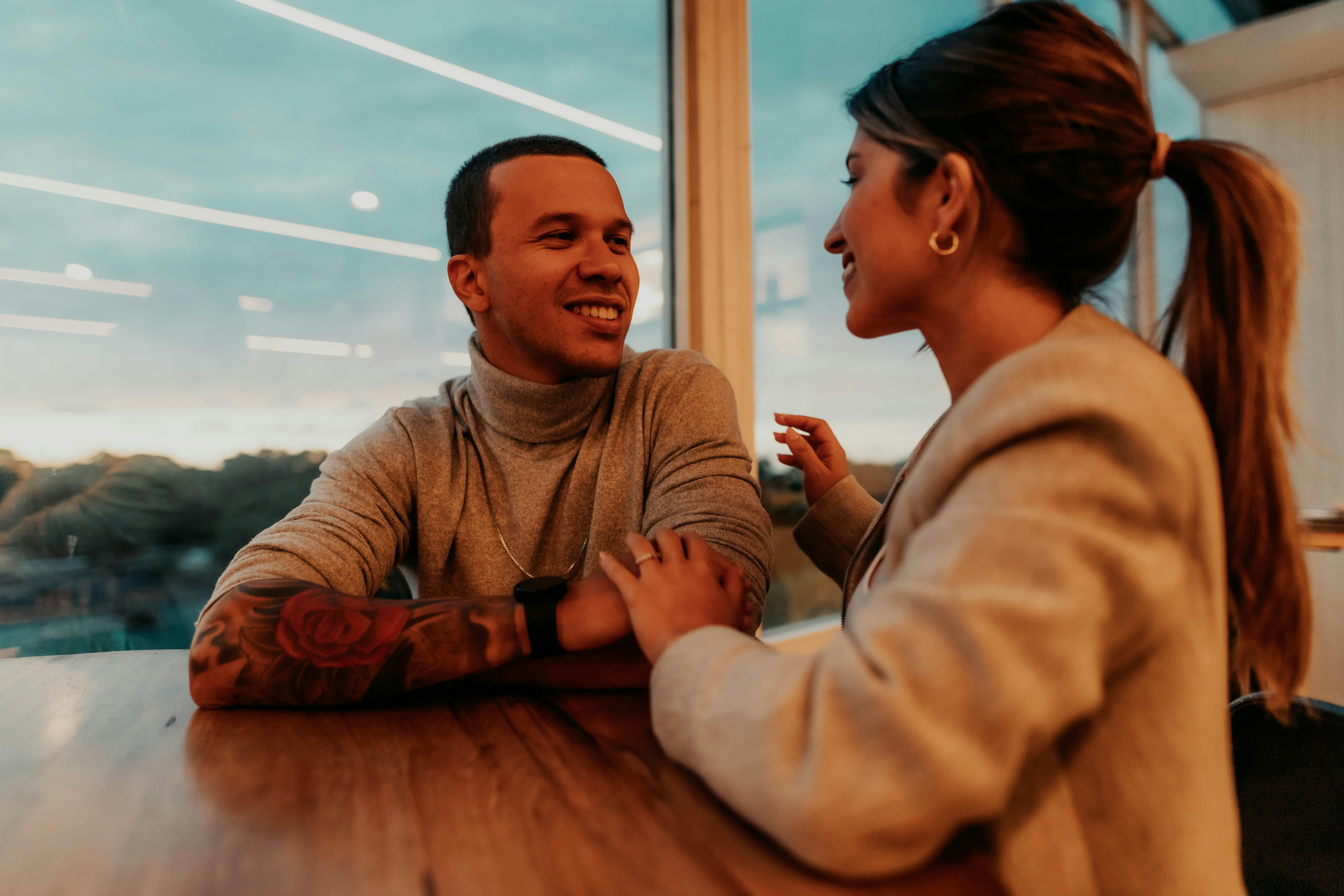 A couple sitting at a table, smiling and enjoying a conversation in a modern setting with large windows.