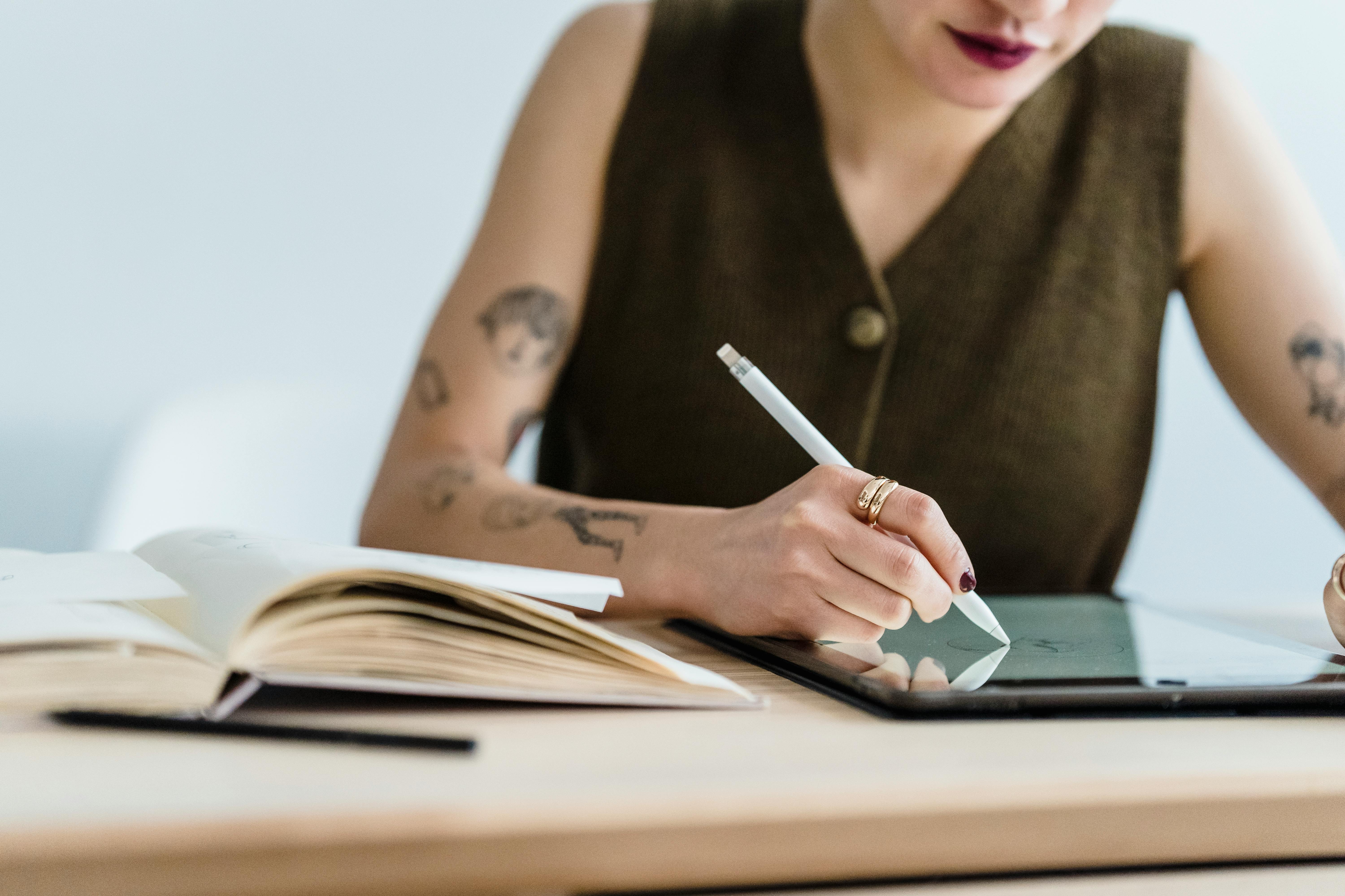 A person using a stylus on a tablet, sketching while sitting at a desk with an open book and a pen.