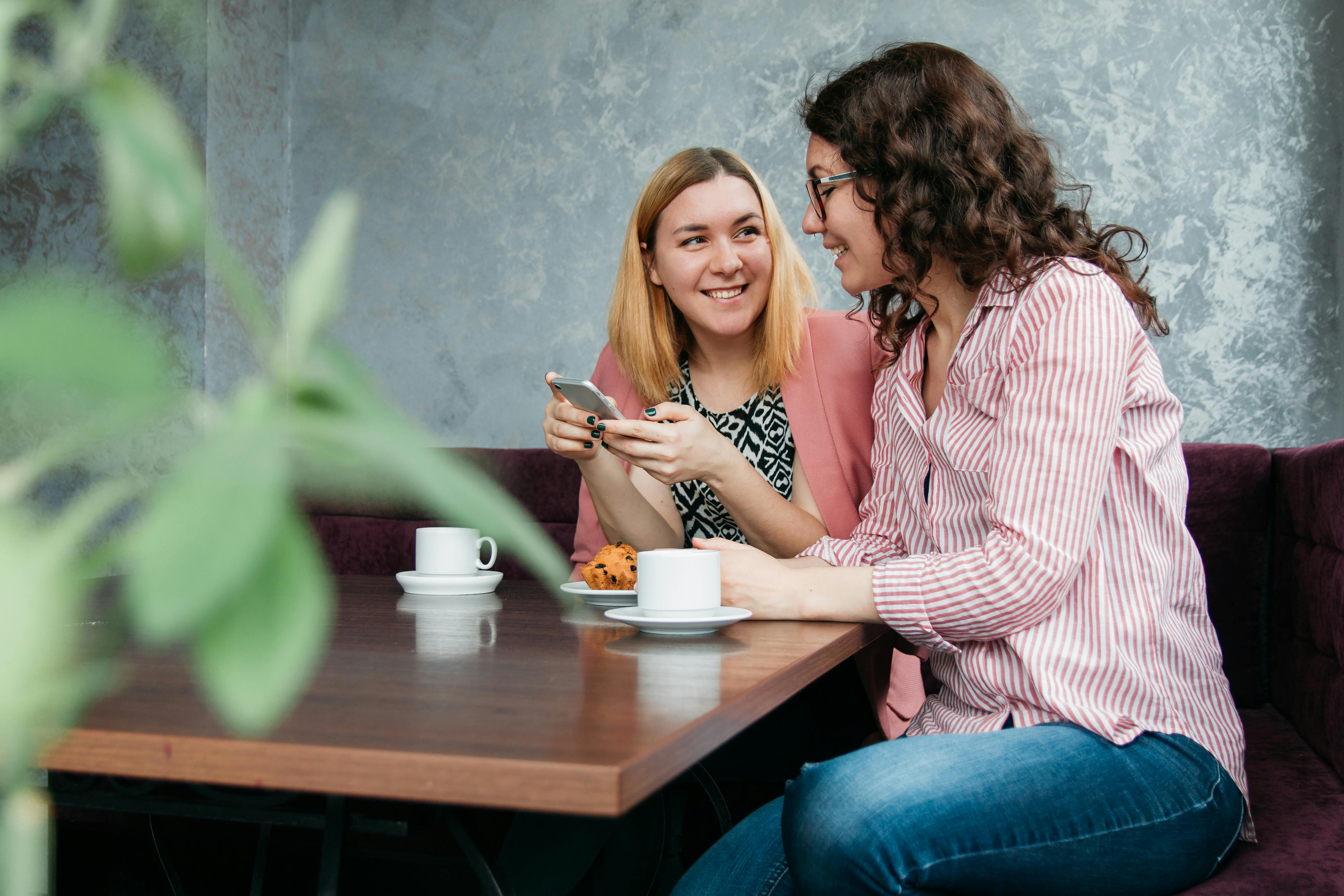 Two women sitting at a café table, smiling and interacting with each other.