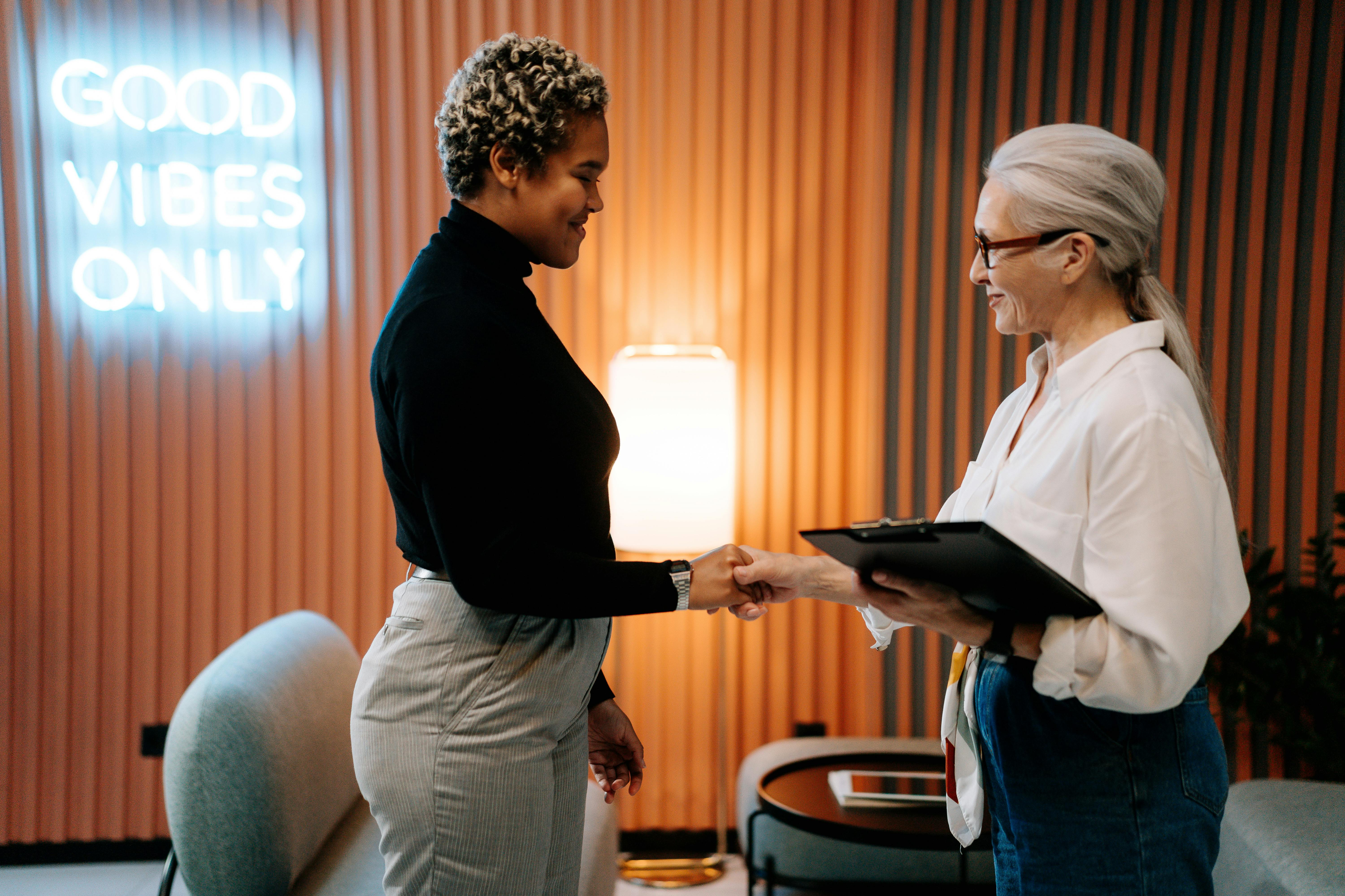 Two women shaking hands in a stylish office environment with a 'Good Vibes Only' neon sign in the background.