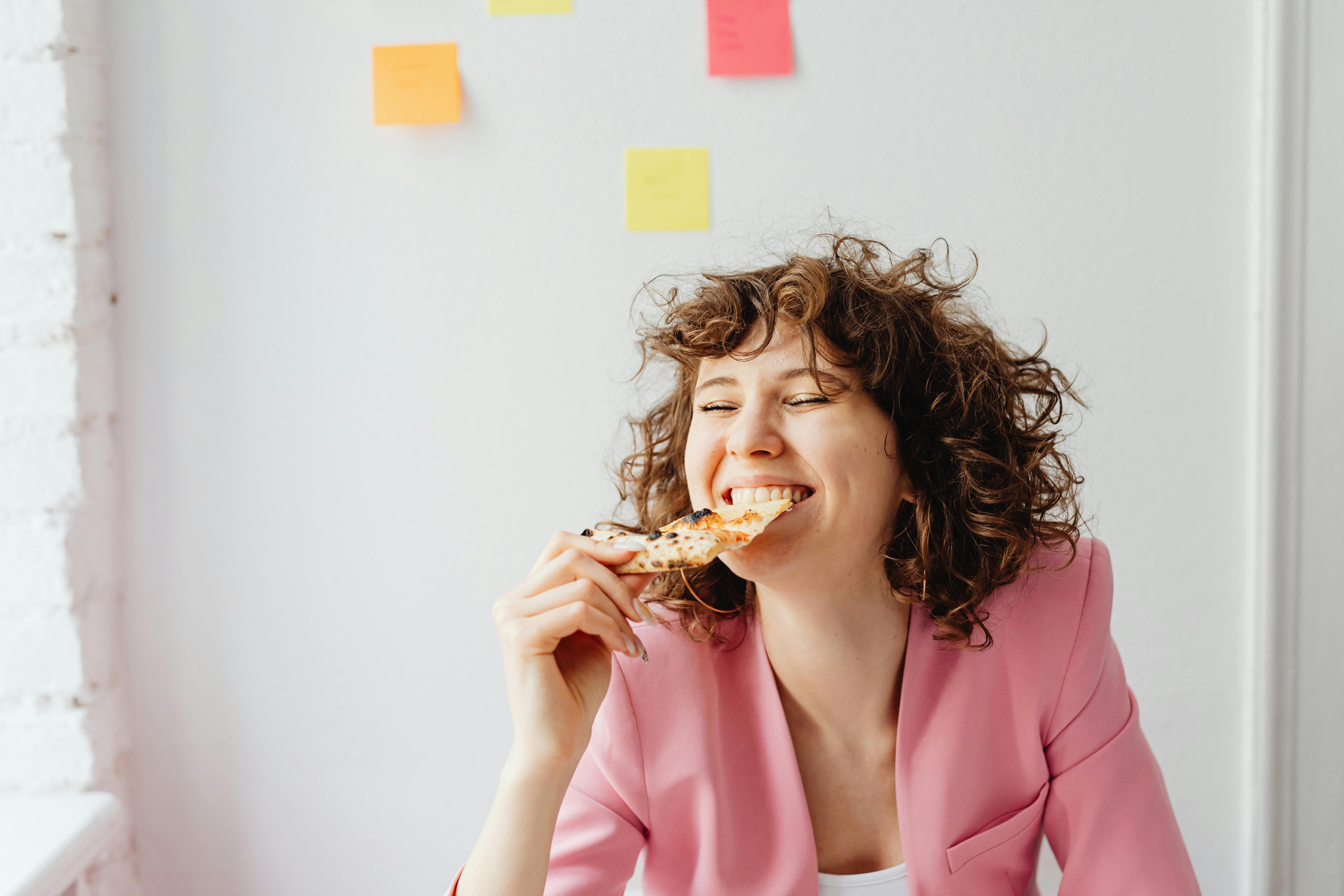 Woman in a pink blazer enjoying a slice of pizza.