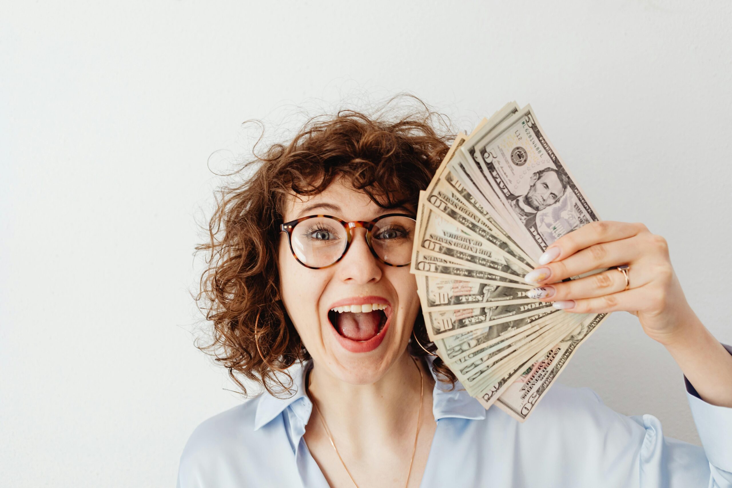 A woman with curly hair wearing glasses is joyfully holding a fan of dollar bills.