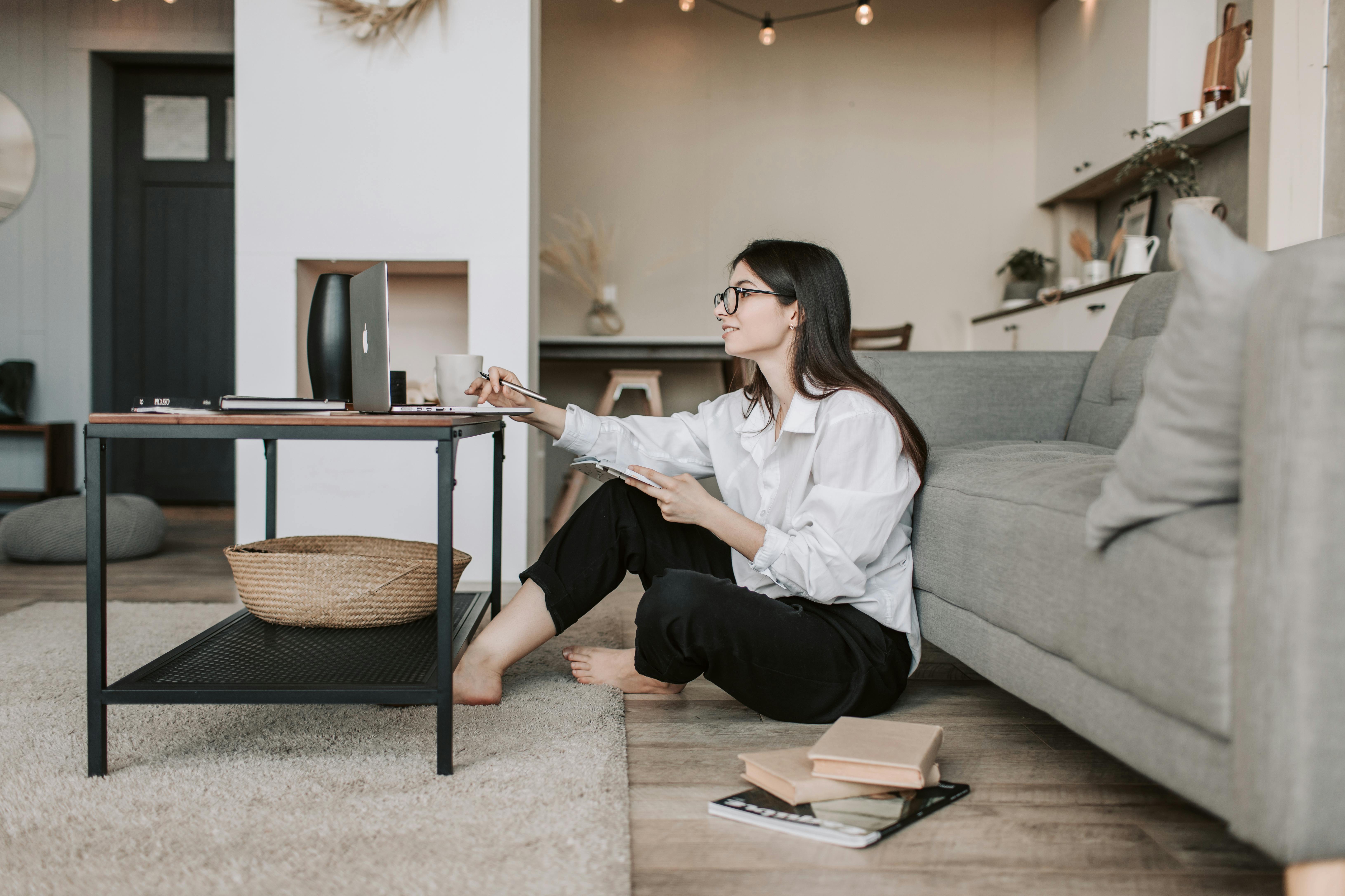 A young woman sitting on the floor beside a coffee table with a laptop, writing in a notebook.