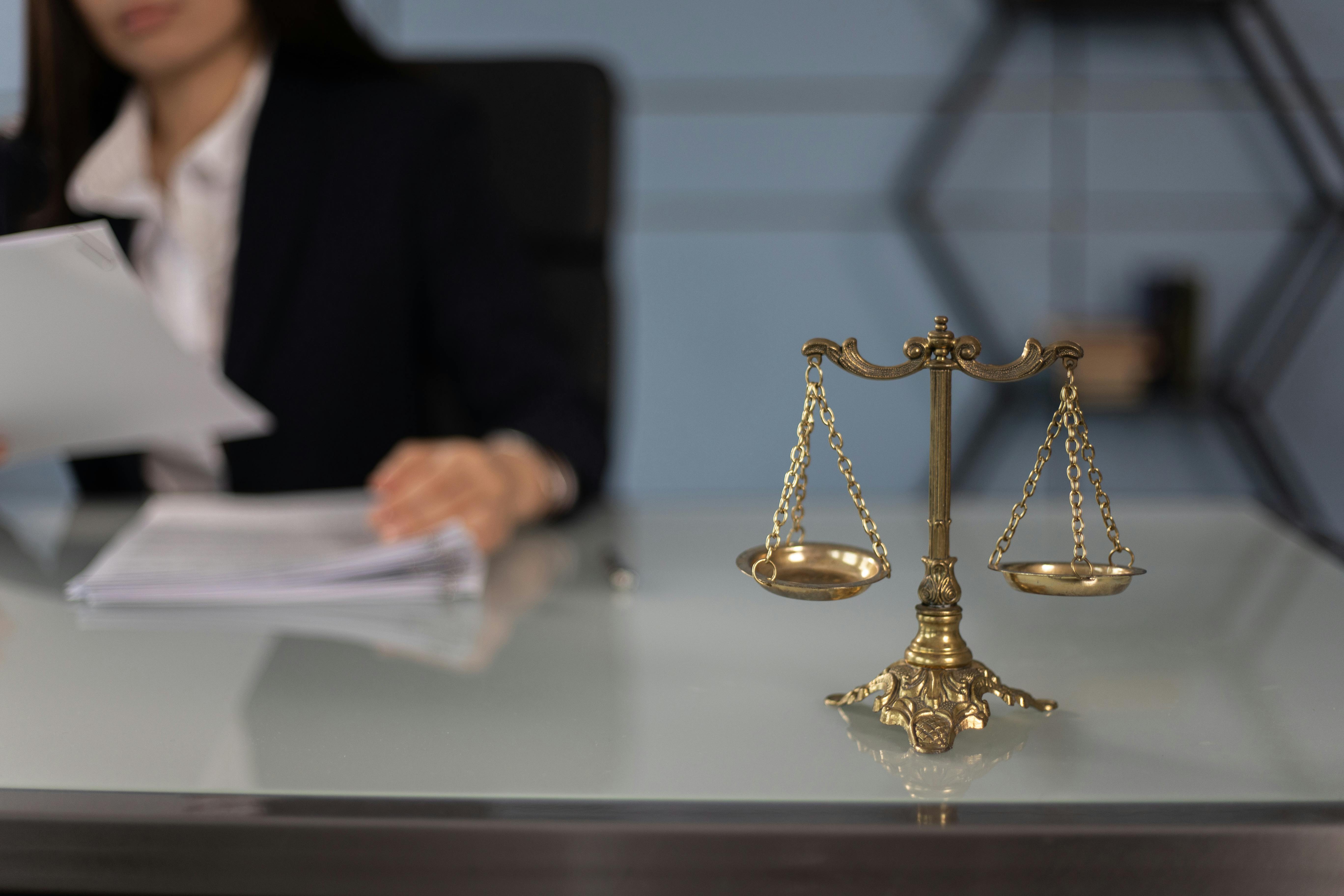 A scale of justice in the foreground with a blurred view of a person holding documents in a professional setting.