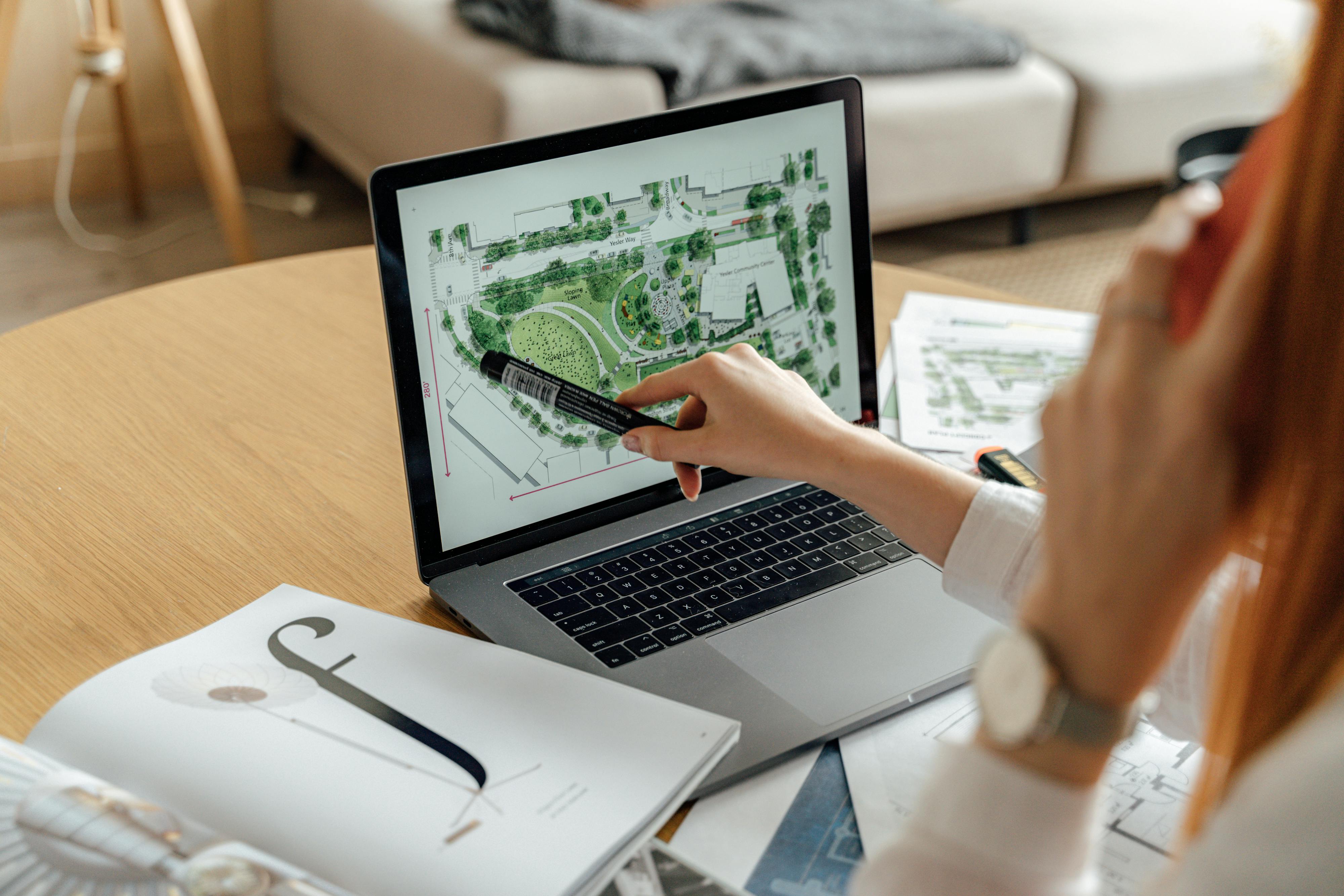 A person analyzing a landscape design on a laptop with a marker in hand, surrounded by design documents.