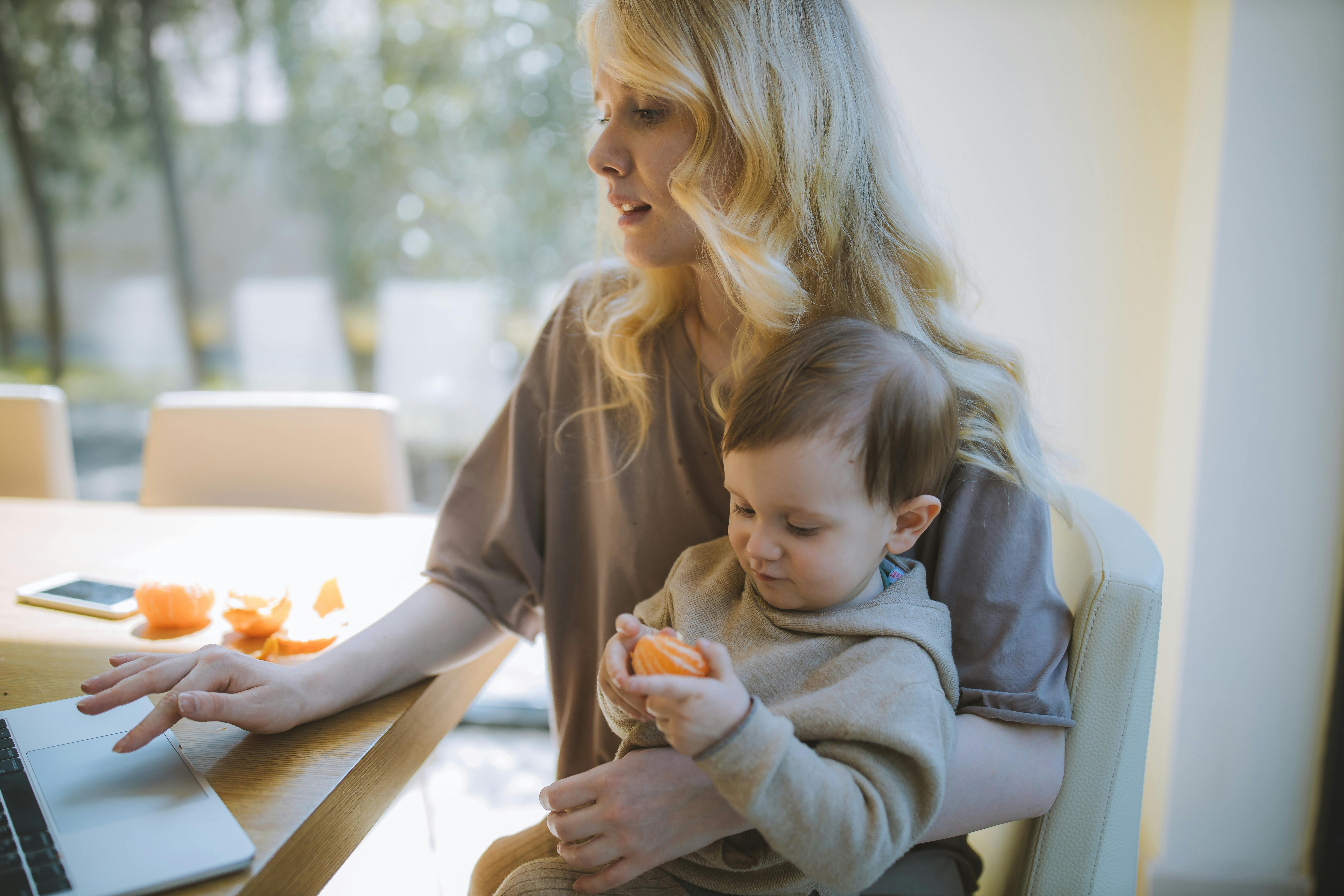 A mother with long blonde hair sits at a table with her toddler, who is holding a small piece of food. They are in a bright, airy room.