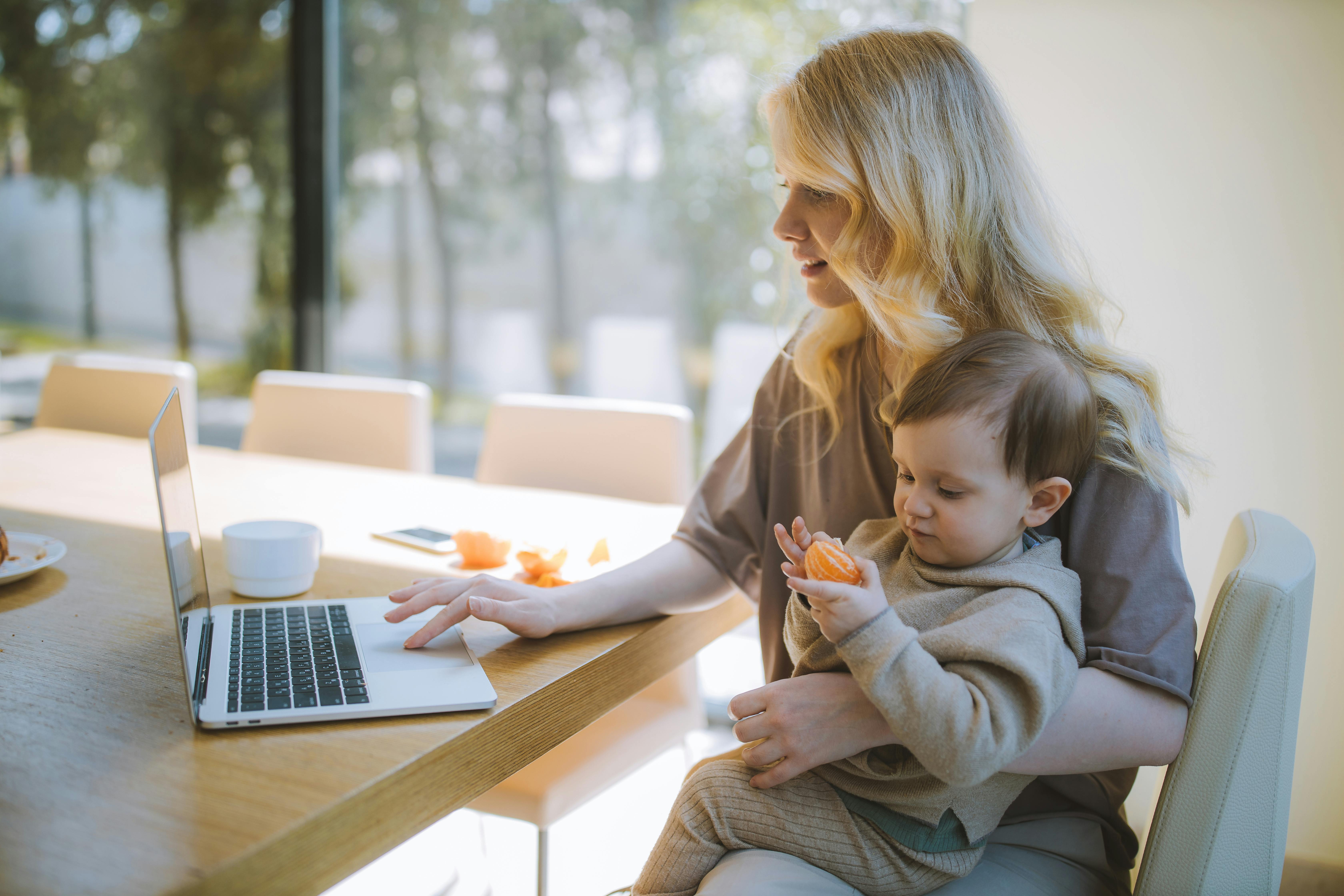 A mother working on a laptop at a table with her baby in her lap, holding a piece of fruit.