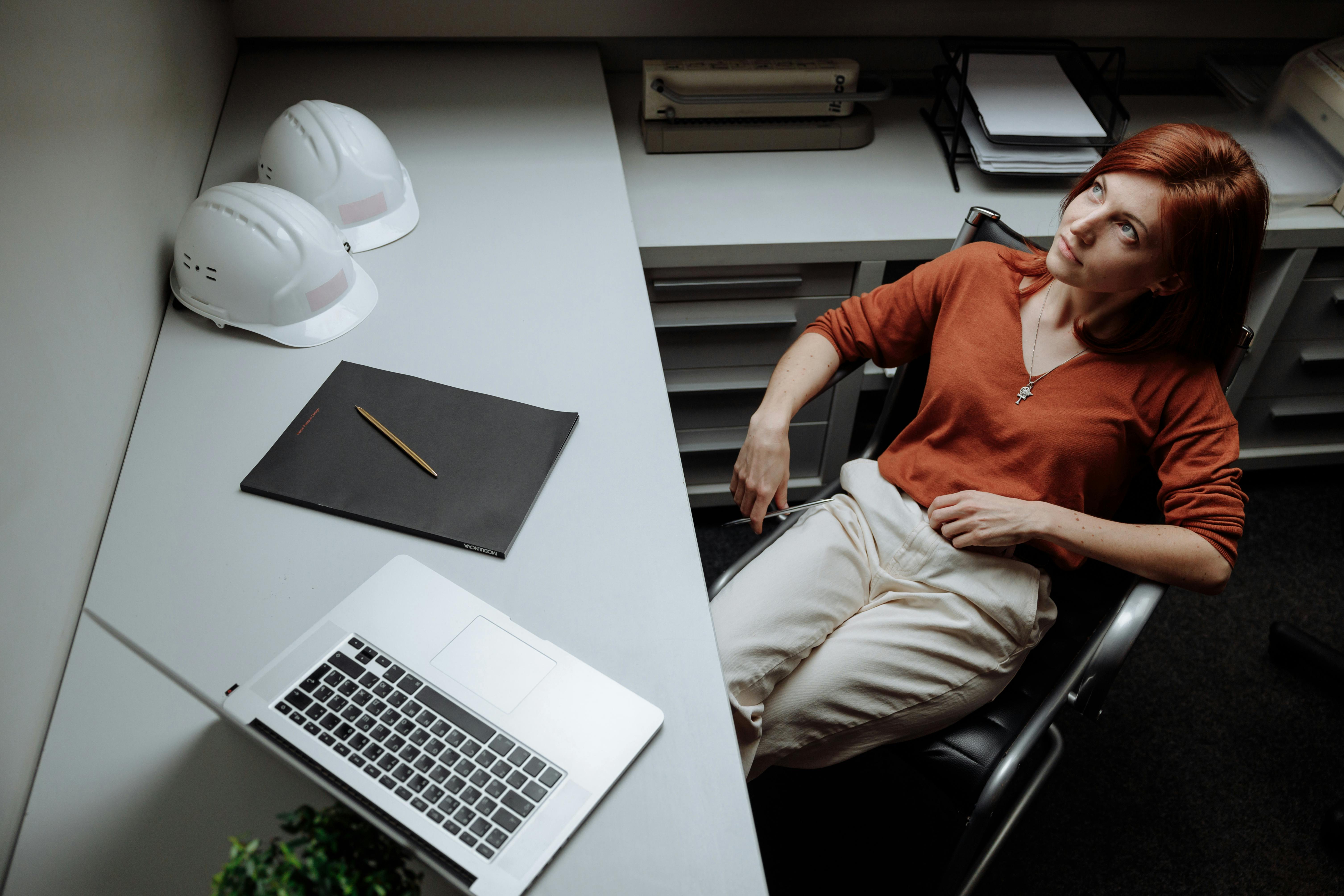 A woman sitting in an office chair beside a desk with construction helmets, a laptop, and a notebook.