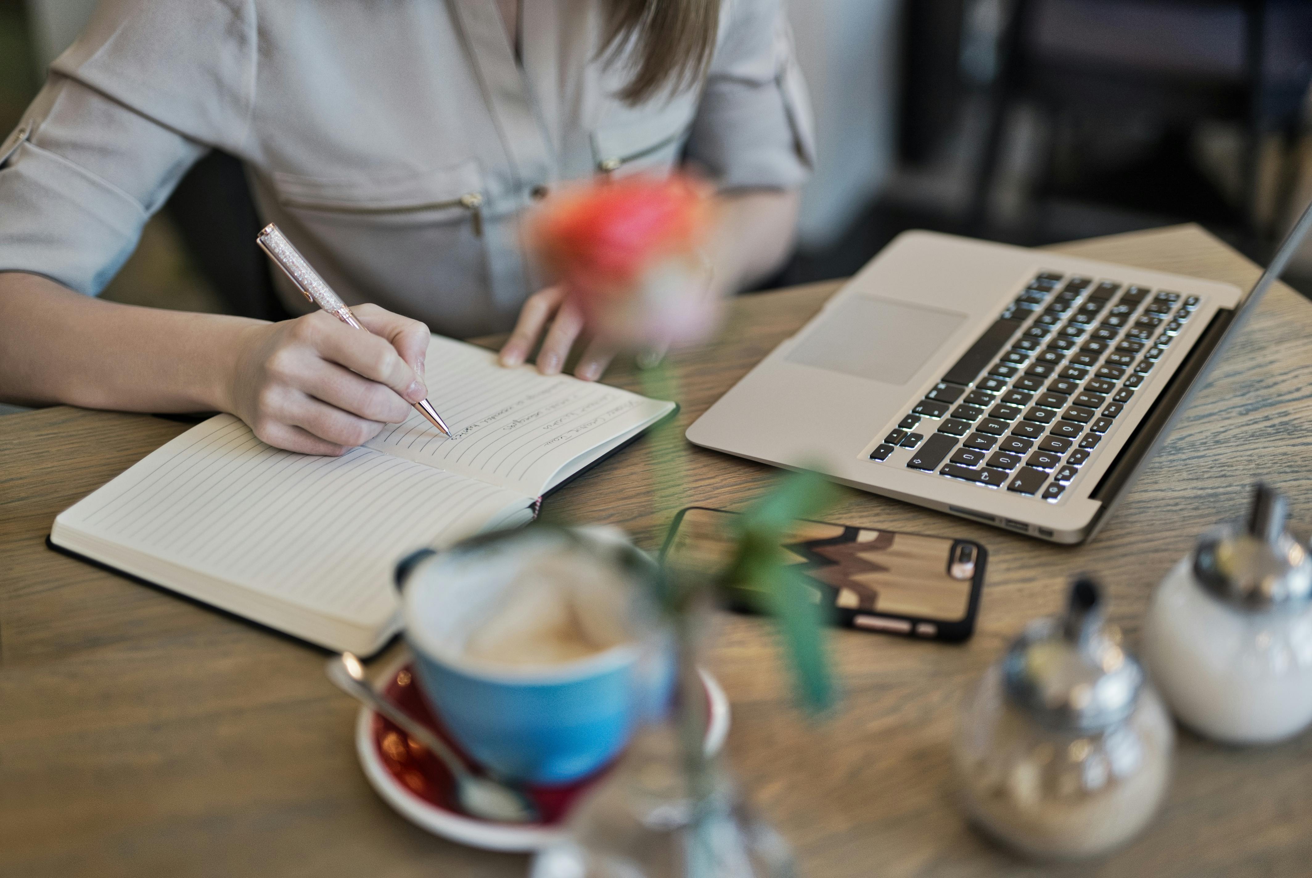 A person writing in a notebook with a laptop and a cup of coffee on the table.