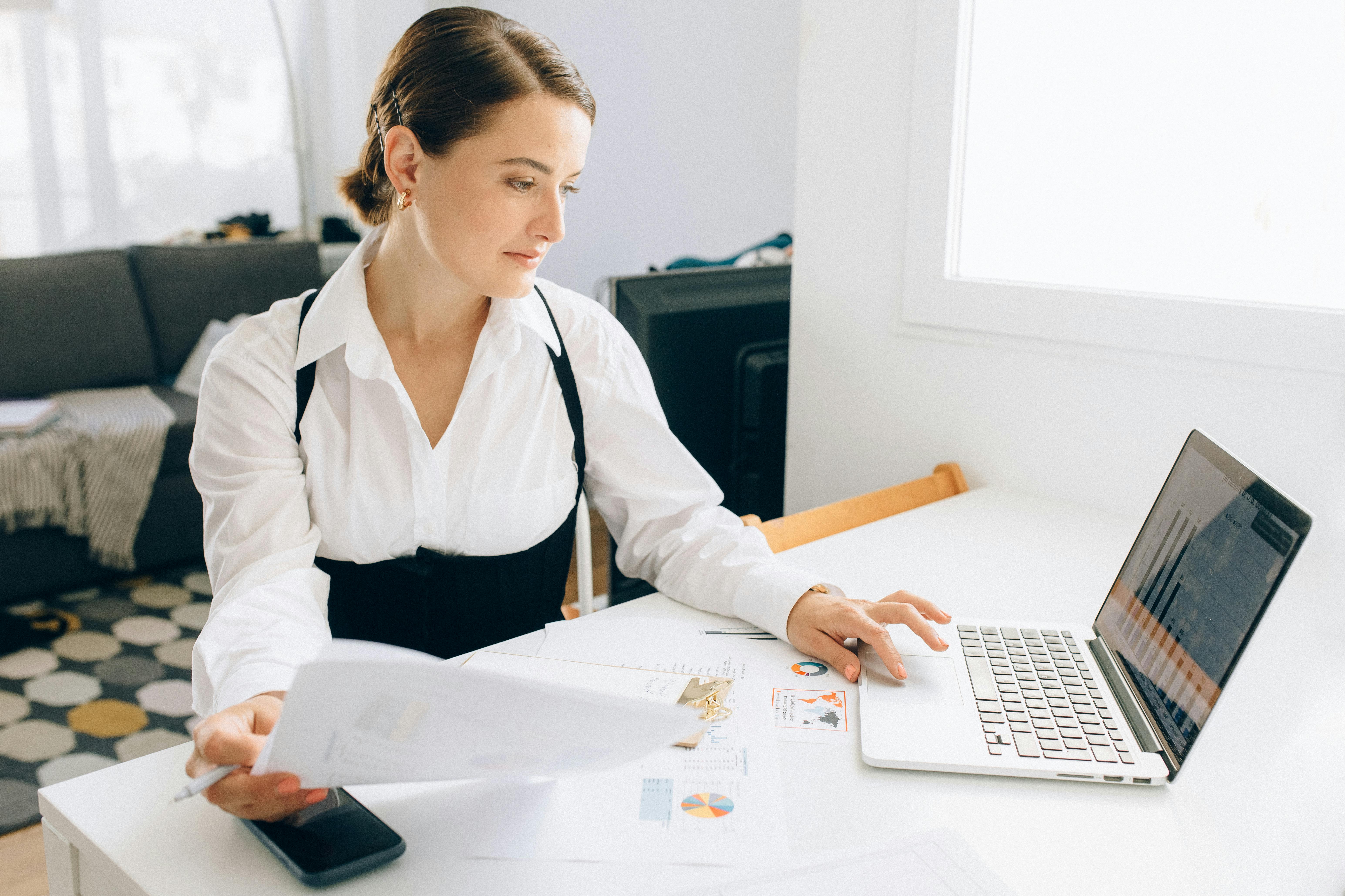 A professional woman in a white shirt, looking at her laptop while holding papers, with a cluttered desk in a modern workspace.