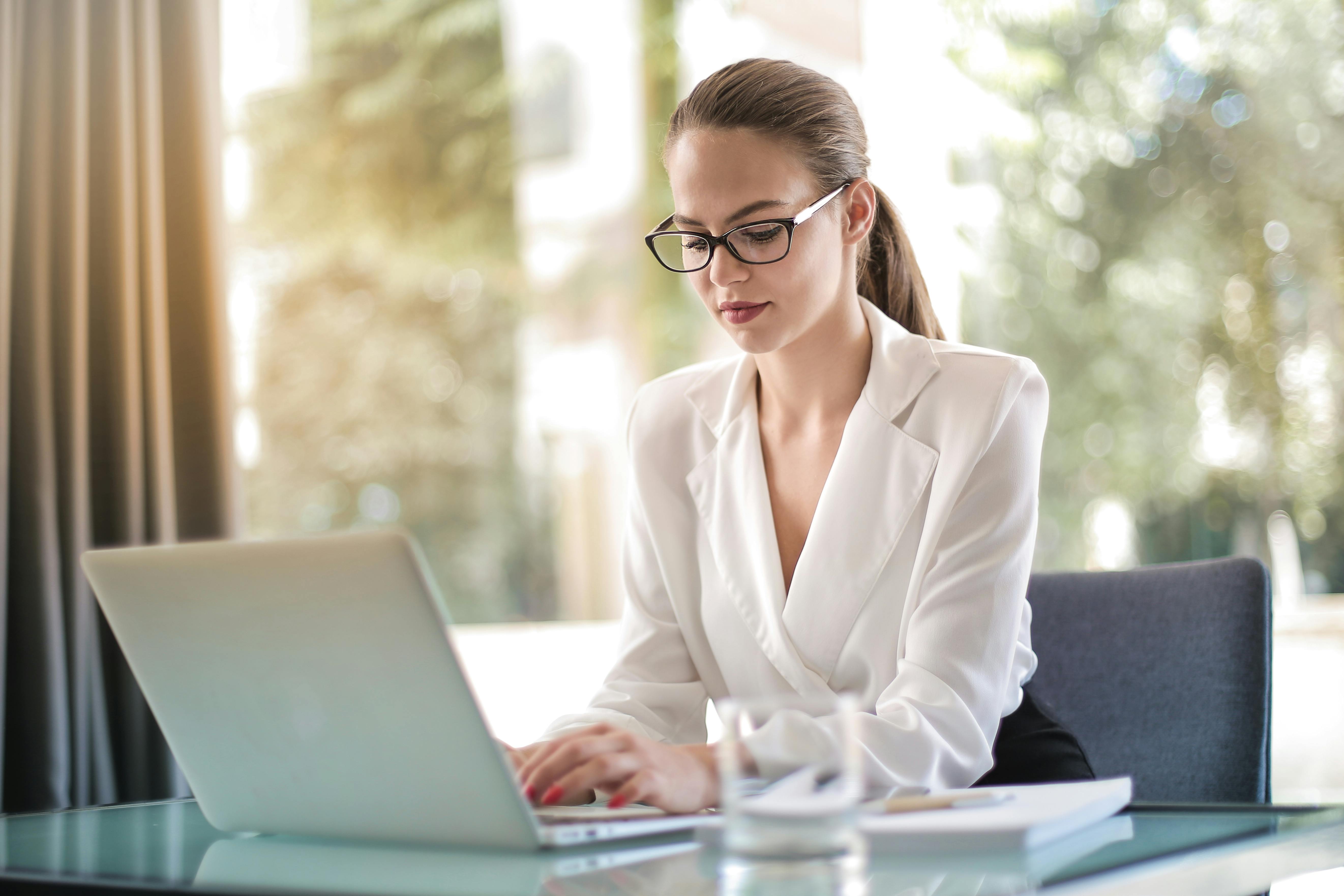 A woman in a white blazer working on a laptop at a table.