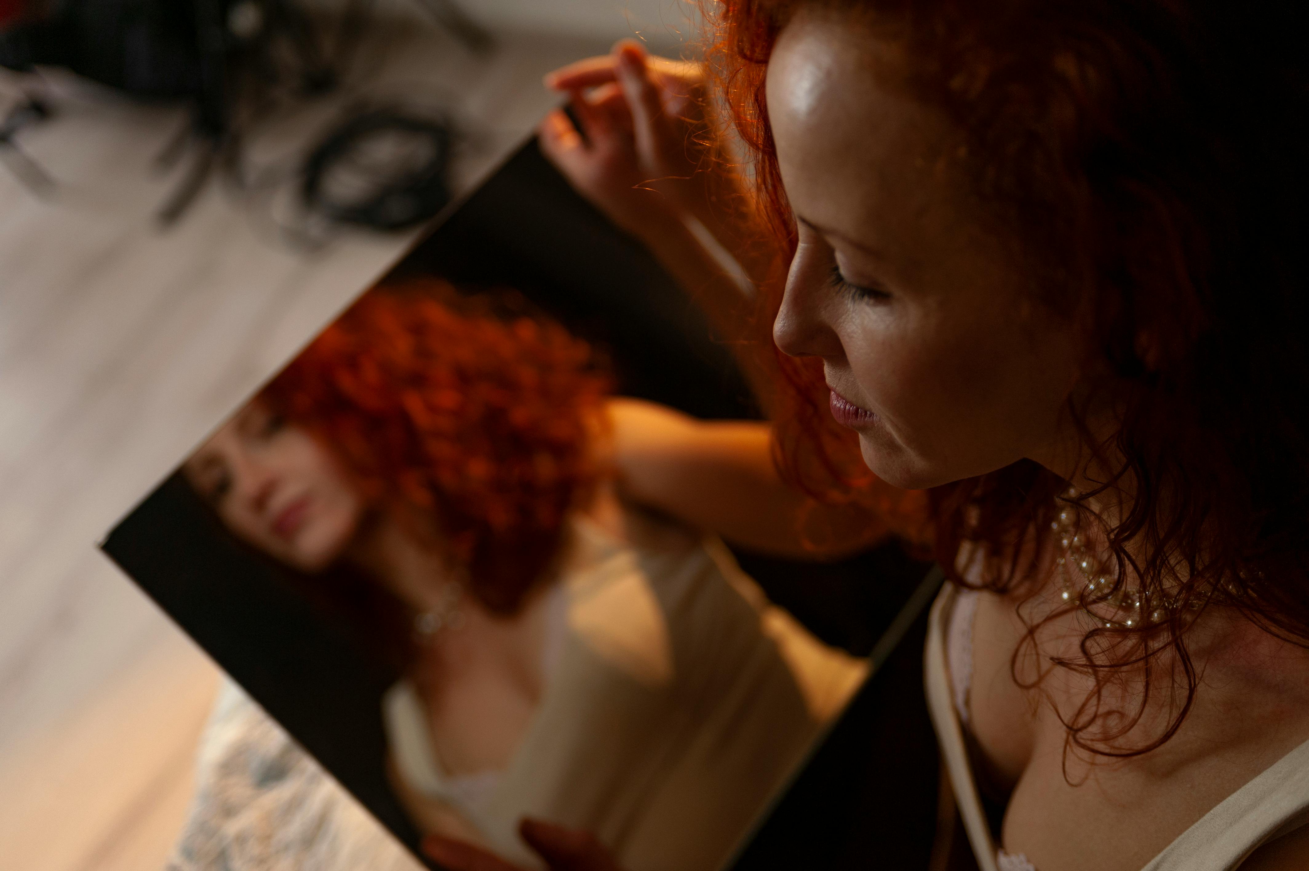 A woman with curly red hair looking into a mirror, softly illuminated, showcasing her profile.