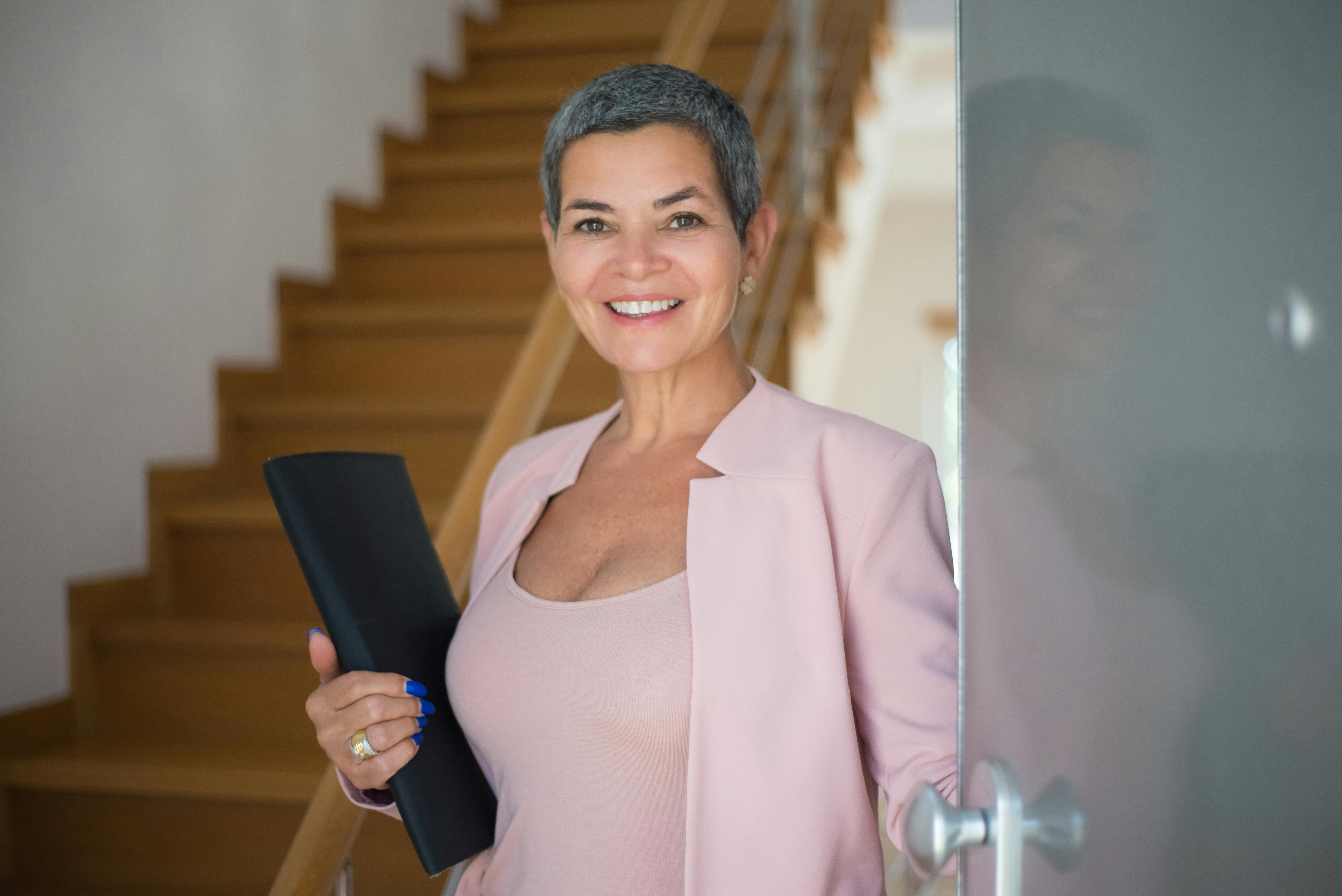 A smiling woman with short hair, wearing a pink blazer, stands by a door holding a black folder.