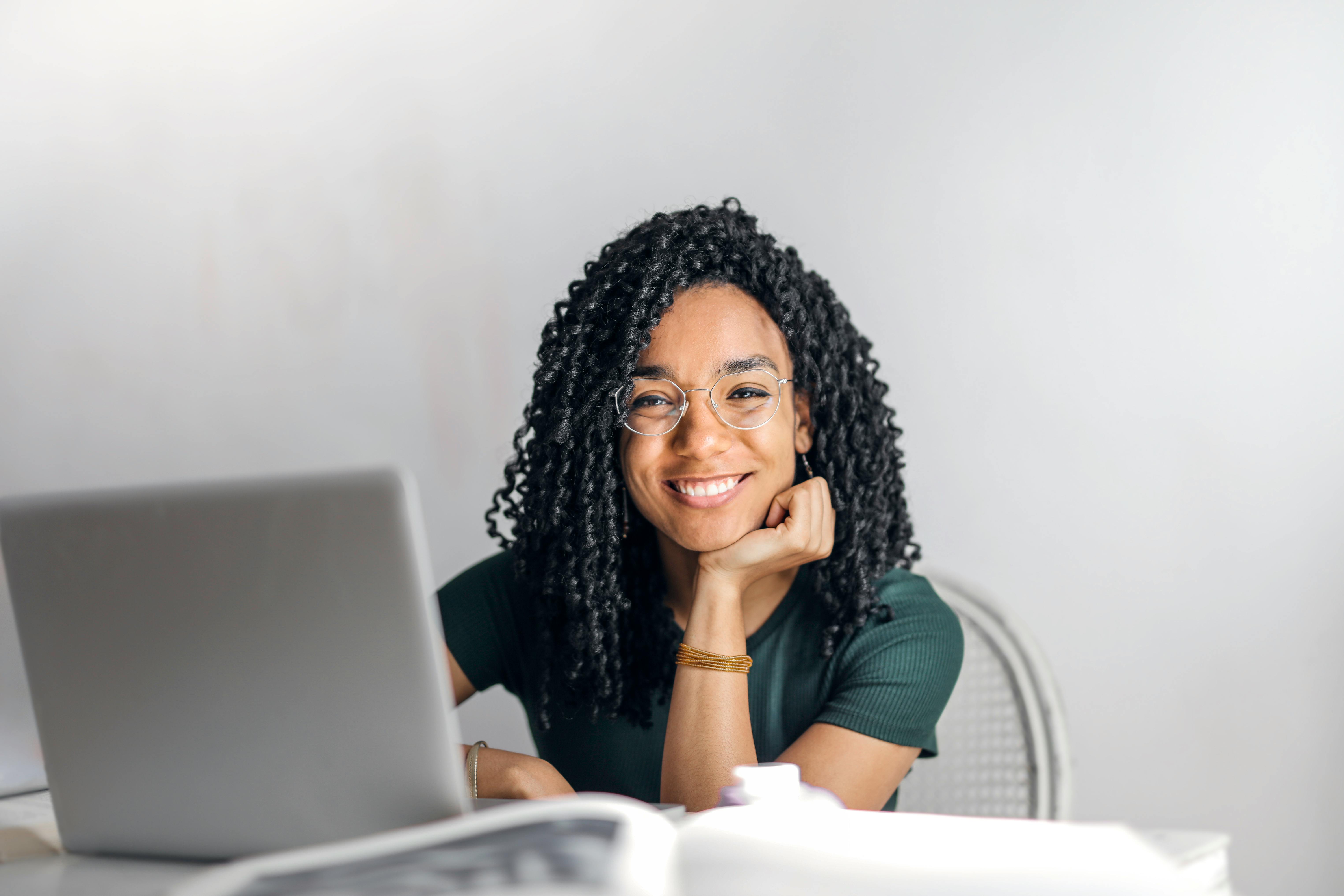 A woman with curly hair and glasses smiling while resting her chin on her hand, sitting in front of a laptop.