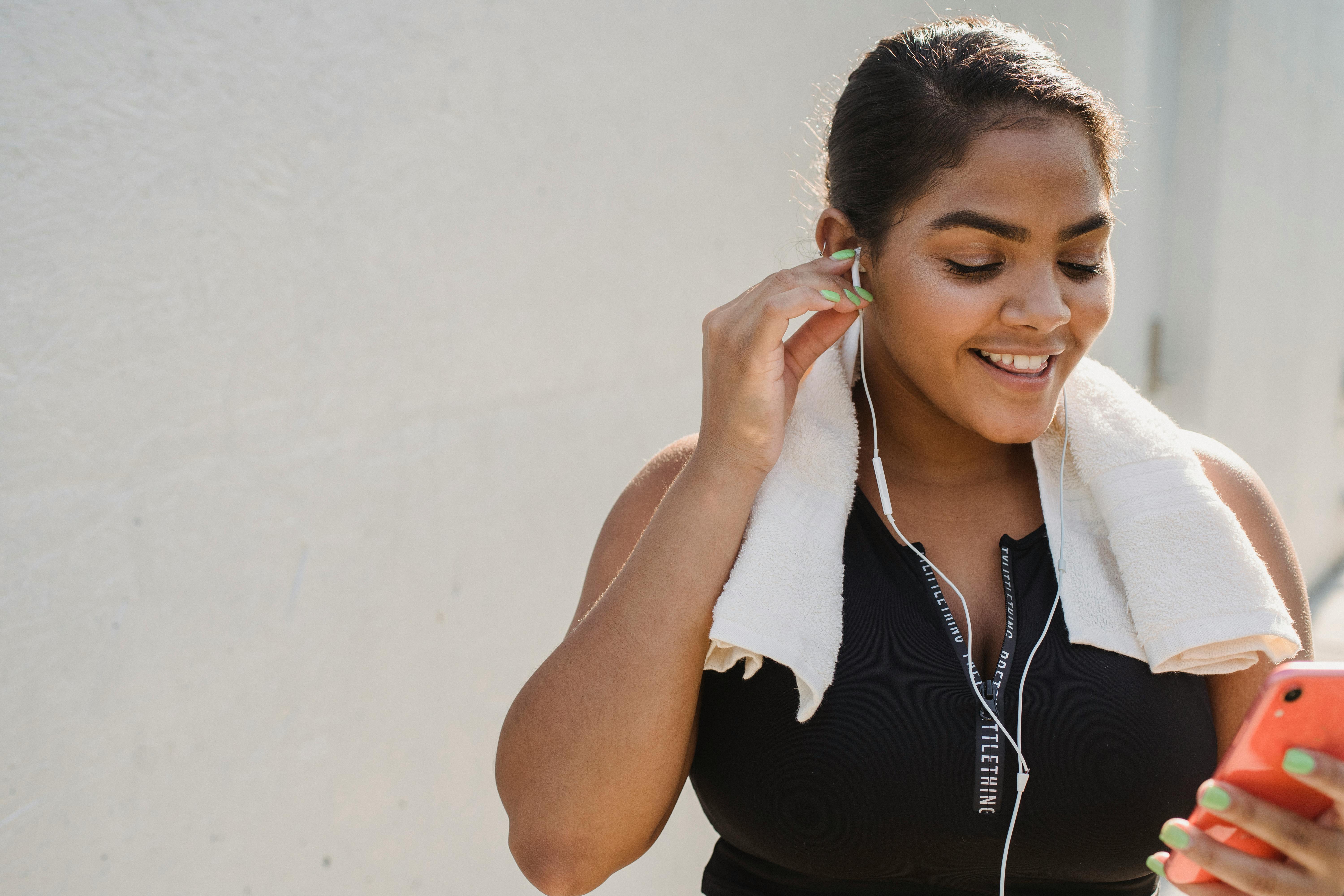 A young woman is smiling and adjusting her headphones while looking at her smartphone.