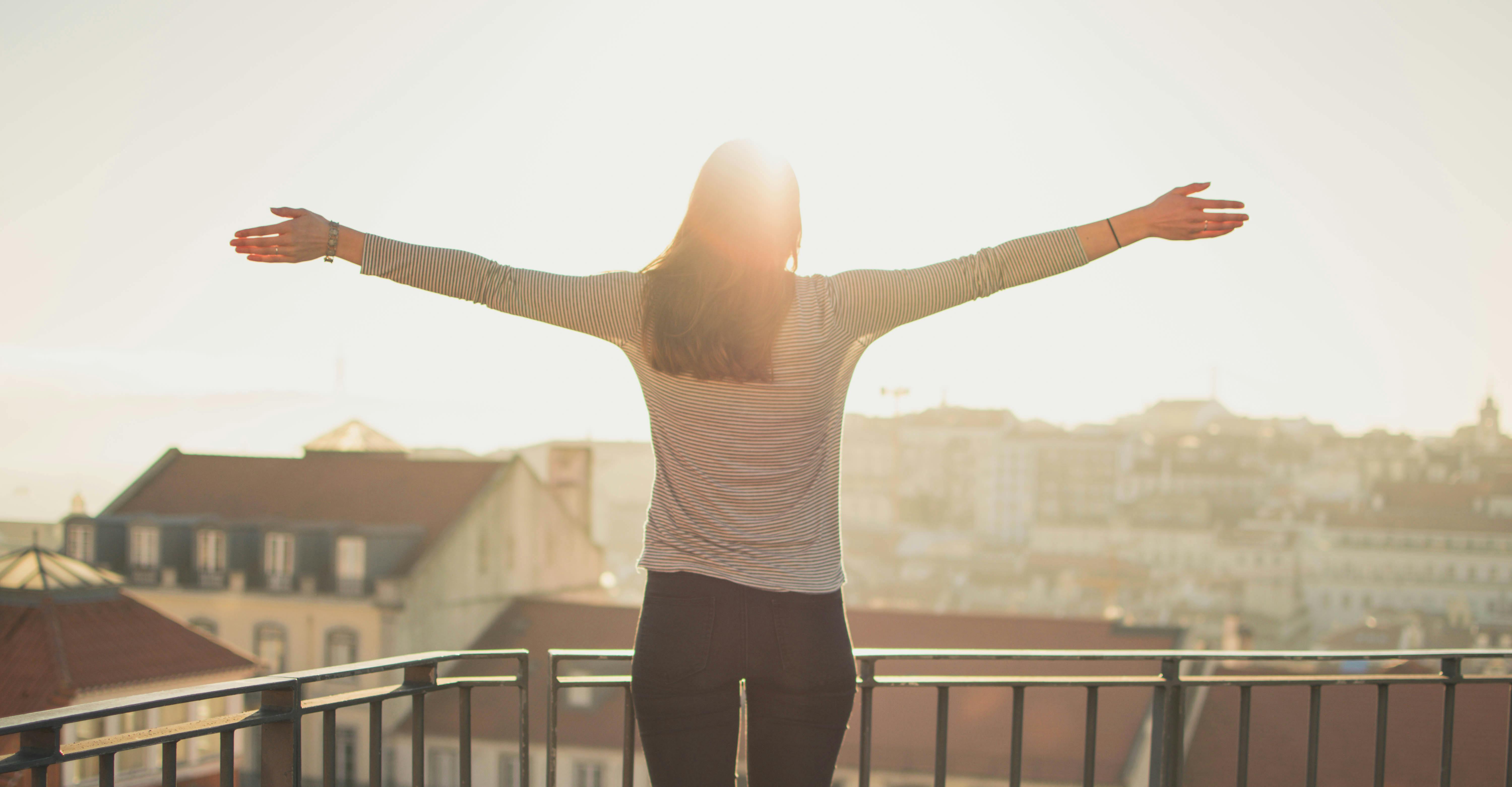 A woman stands with her arms outstretched, enjoying a sunset view over a cityscape.