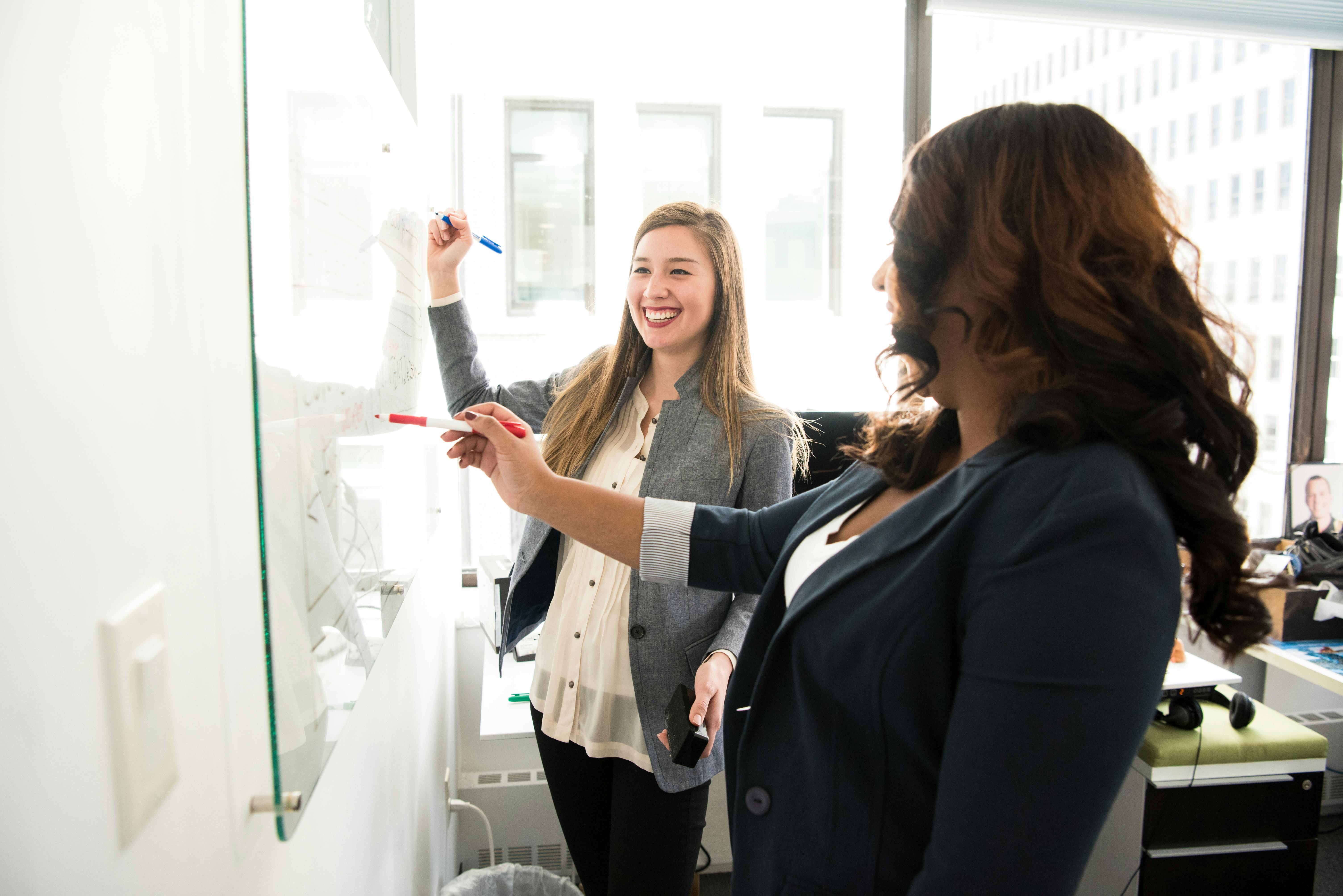 Two women participating in a brainstorming session, writing on a glass board.