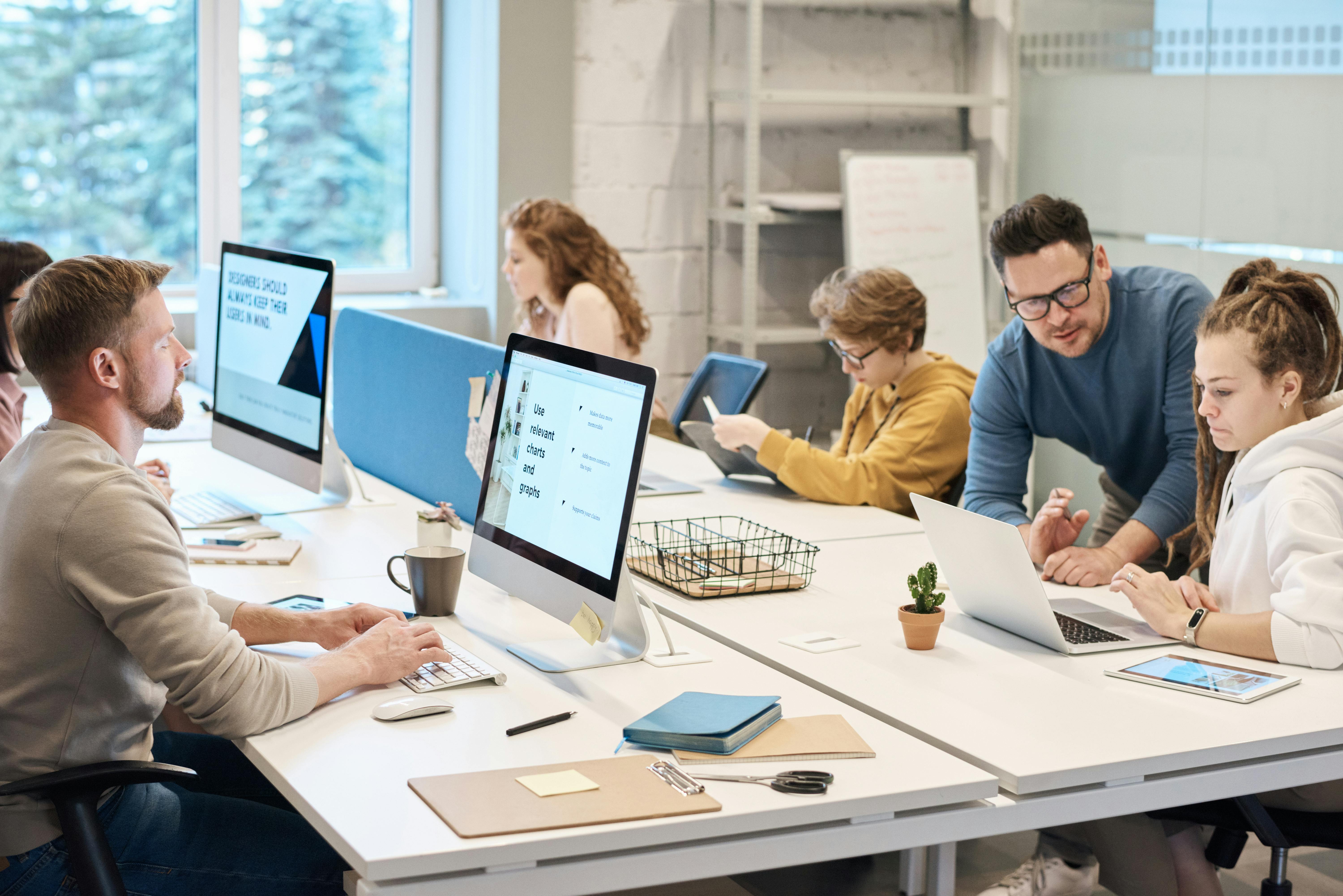 A group of people collaborating in a bright office space with computers and notepads.