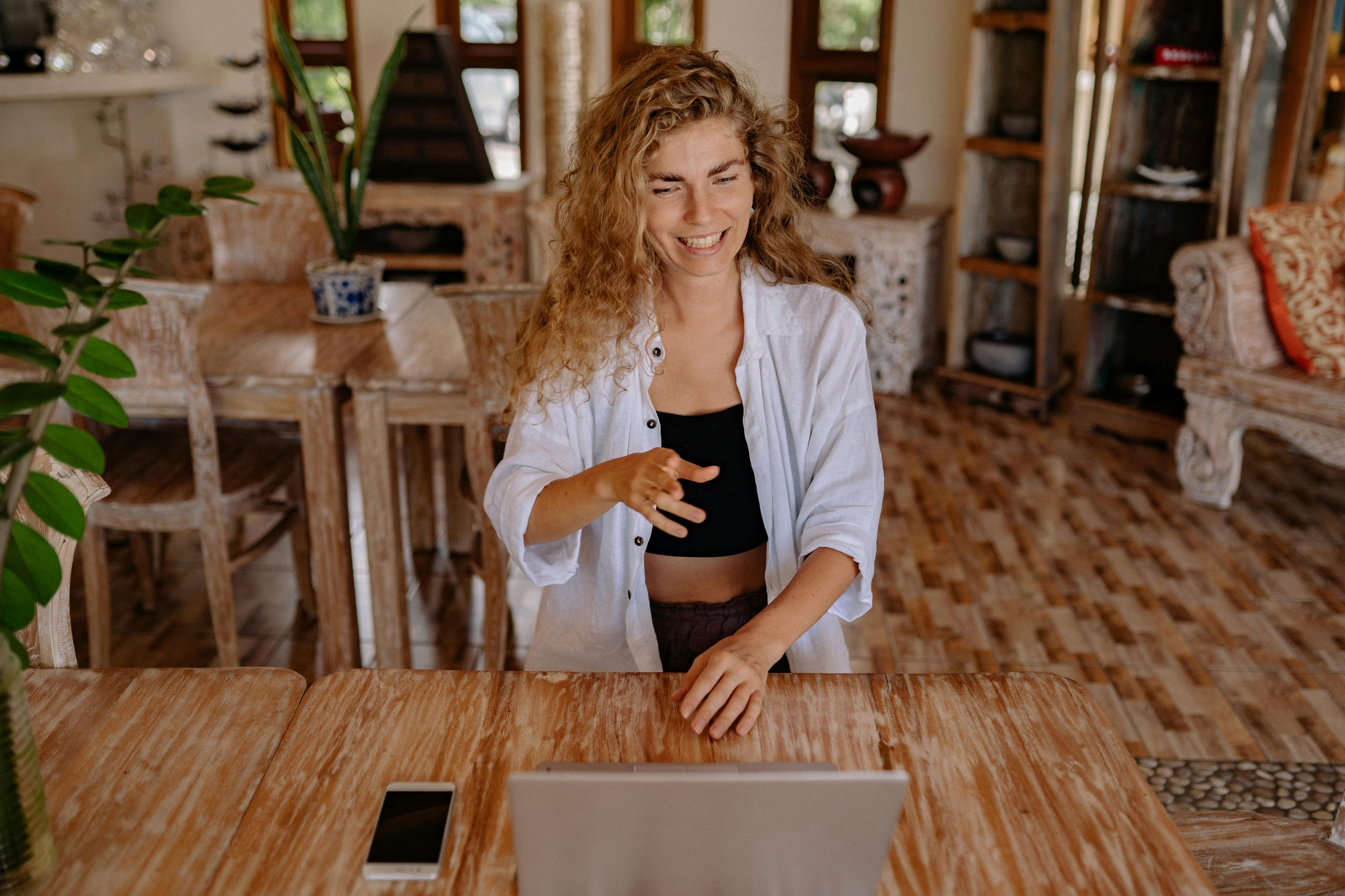 Woman sitting at a wooden table, smiling and gesturing while using a laptop.