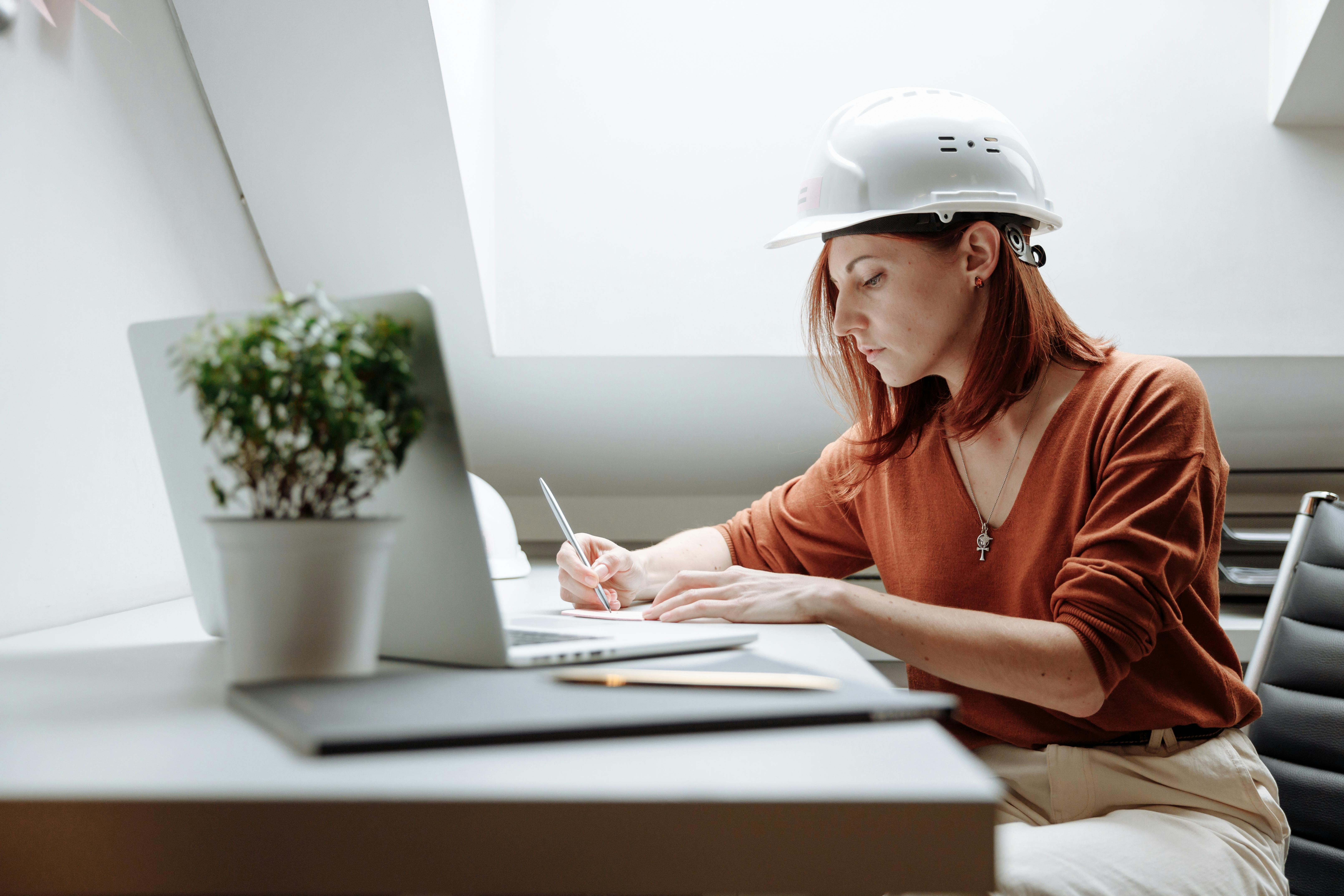 A woman wearing a hard hat sitting at a desk, drawing with a pencil while looking at a laptop.