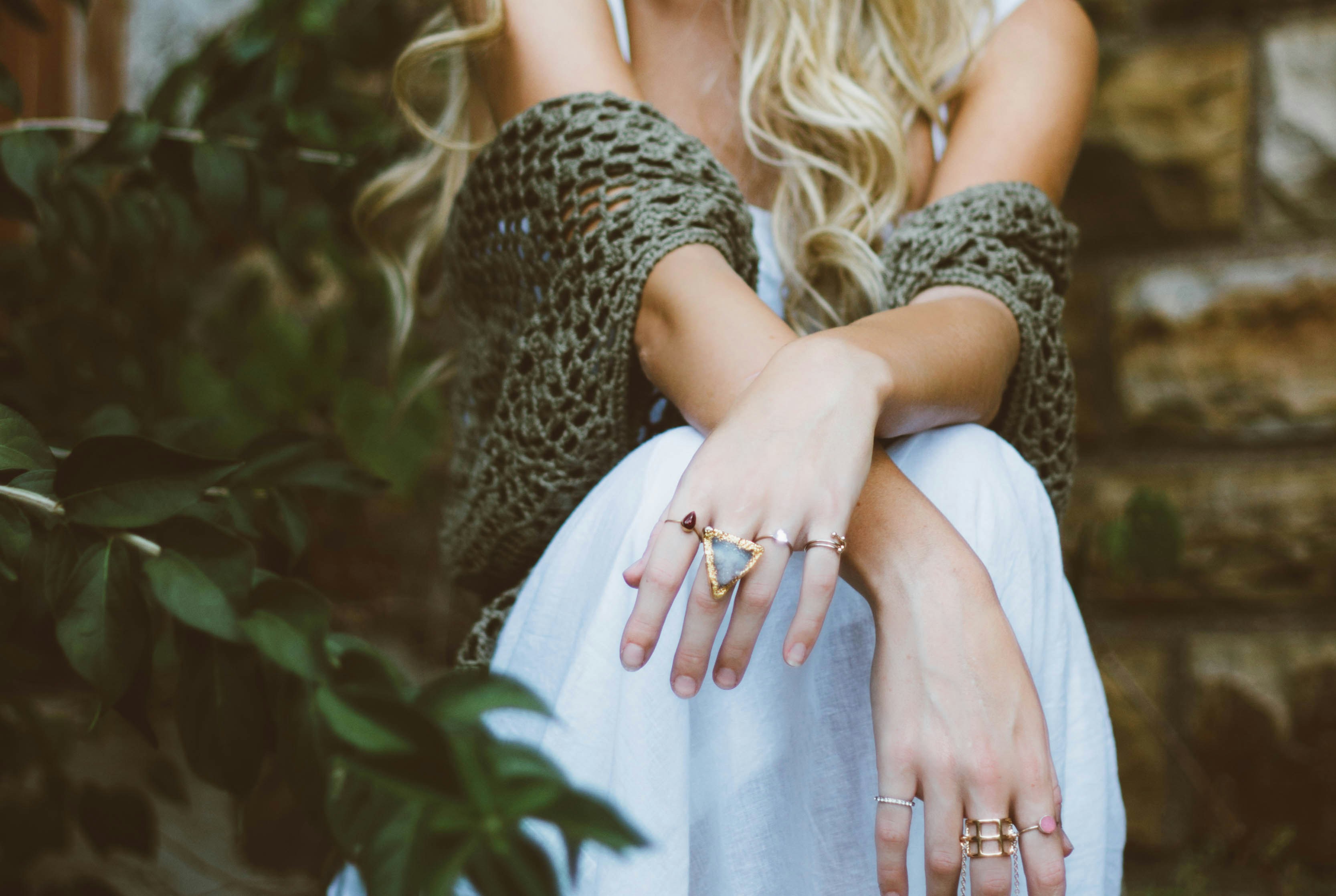 Close-up of a woman's hands adorned with rings, wearing a white dress, sitting near greenery.