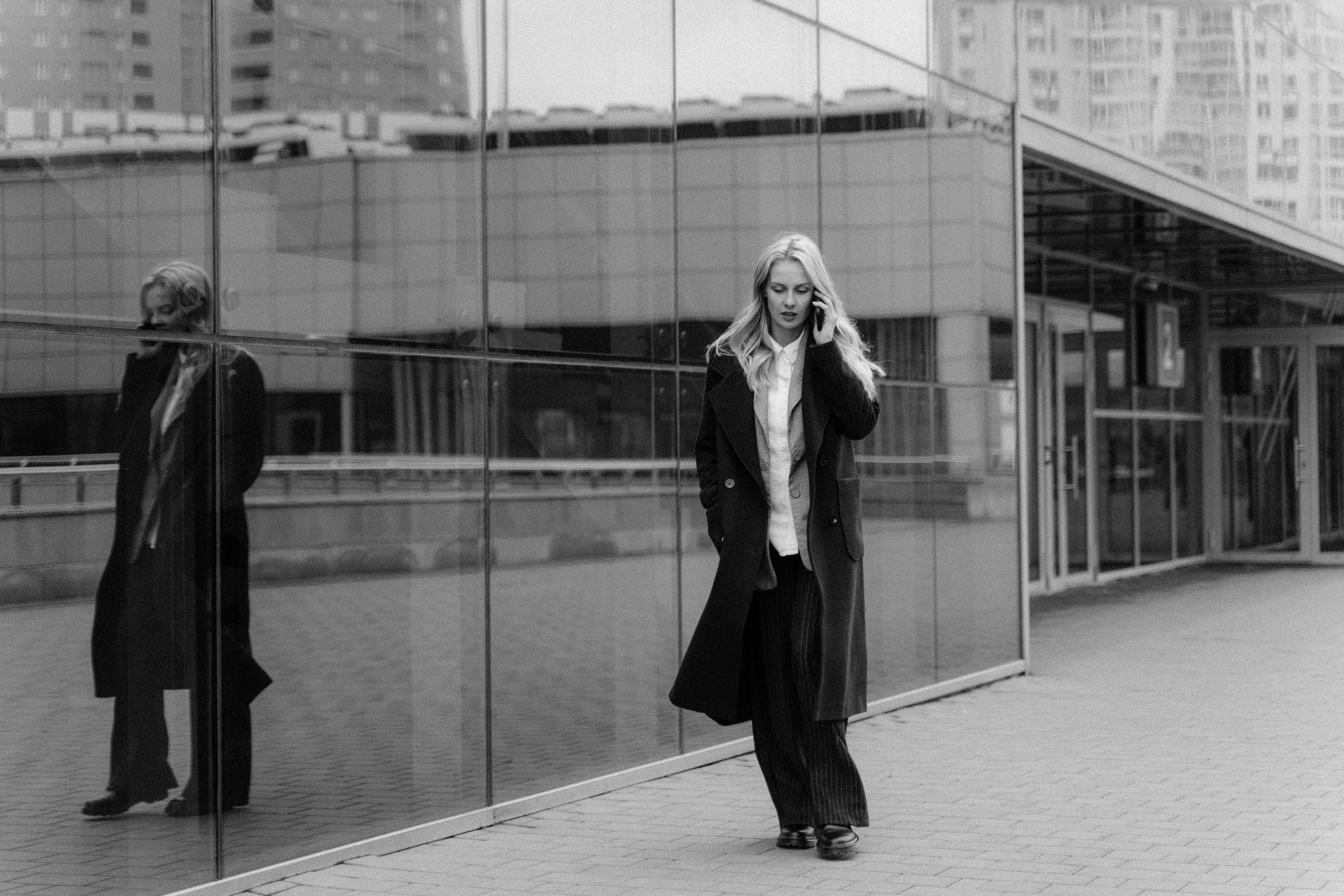 A woman in a black coat talking on her phone with a reflection in a glass wall.