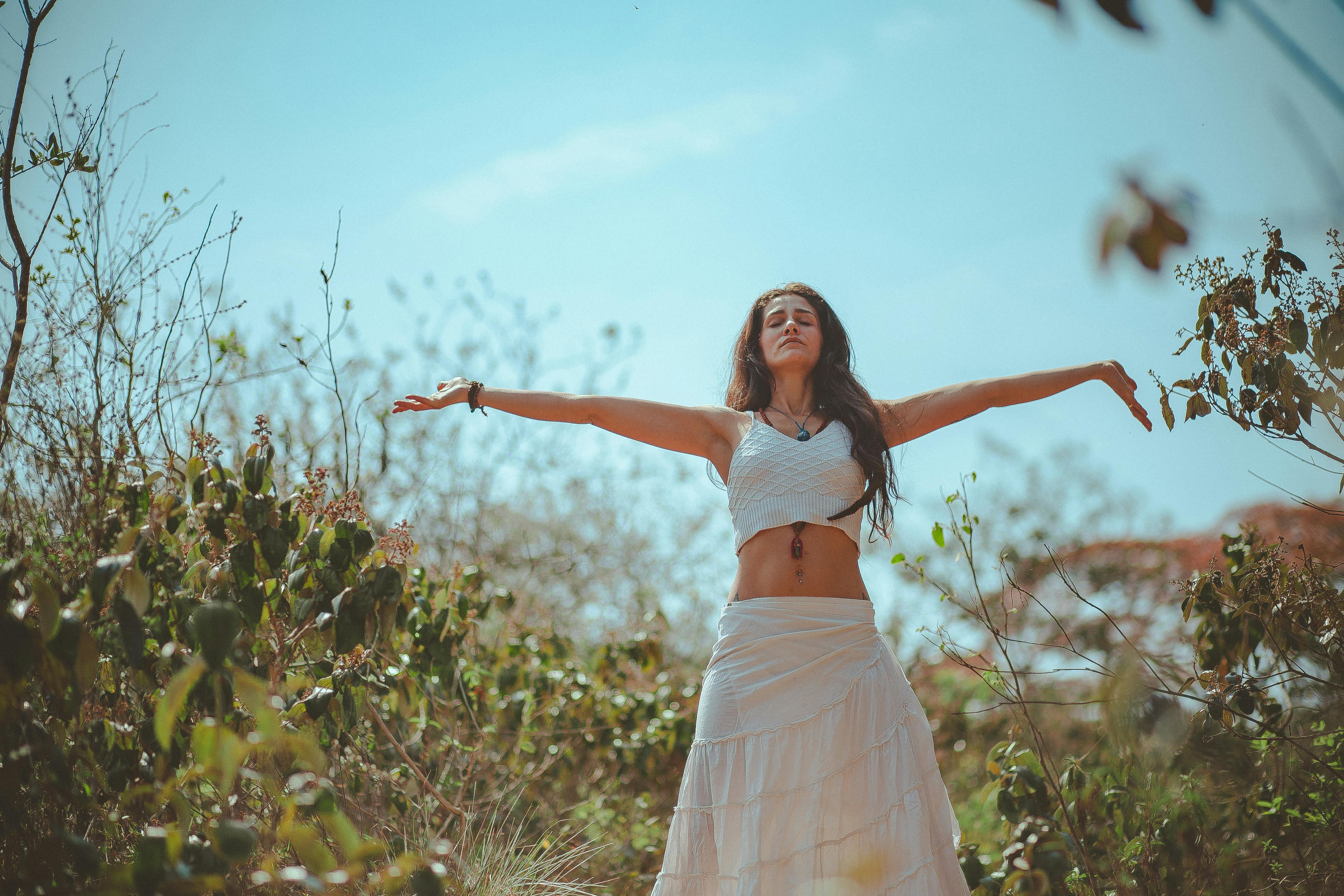 A woman with long hair standing in a natural setting, arms outstretched towards the sky.