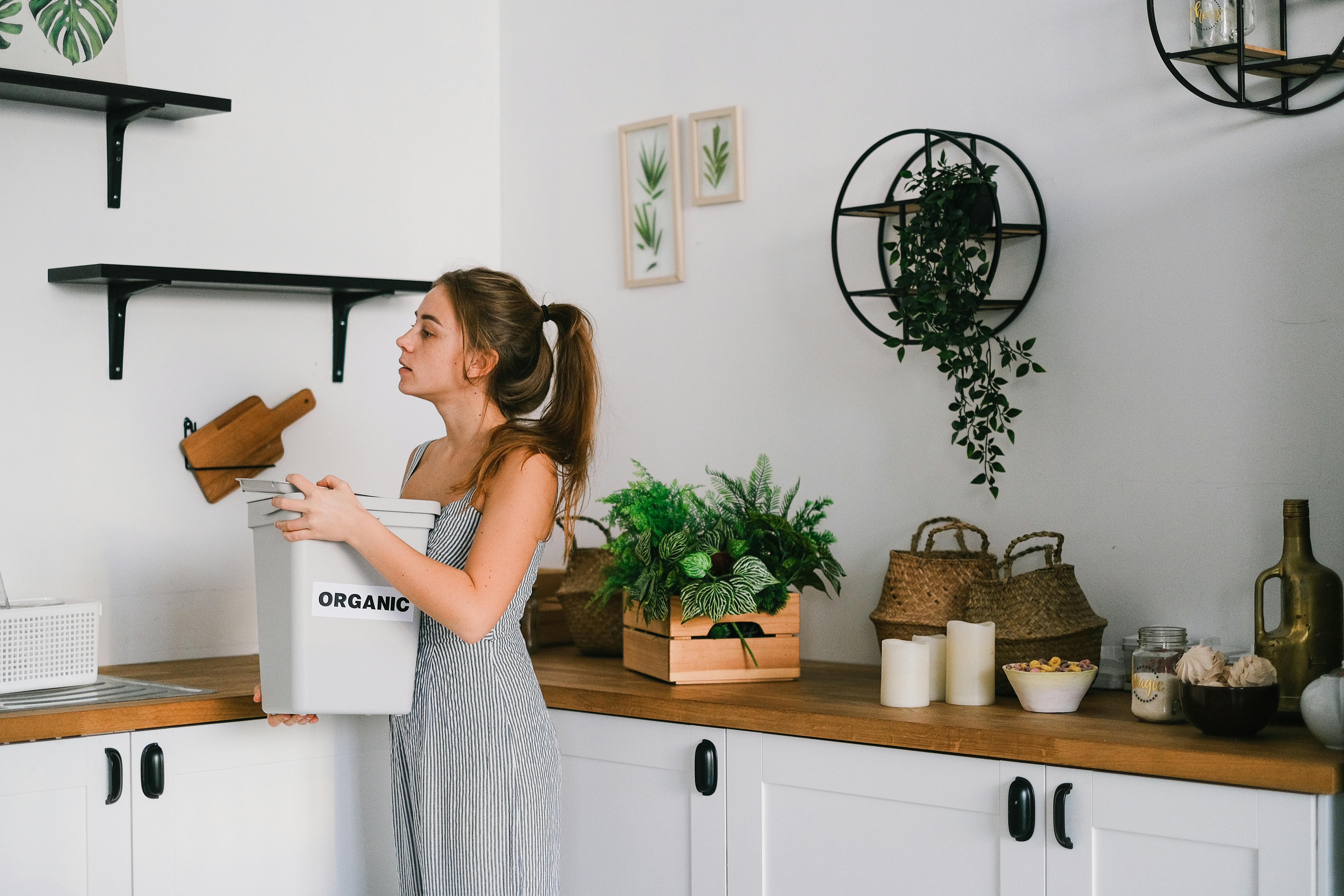 A woman holding an organic waste bucket in a bright kitchen with plants and decorative items.