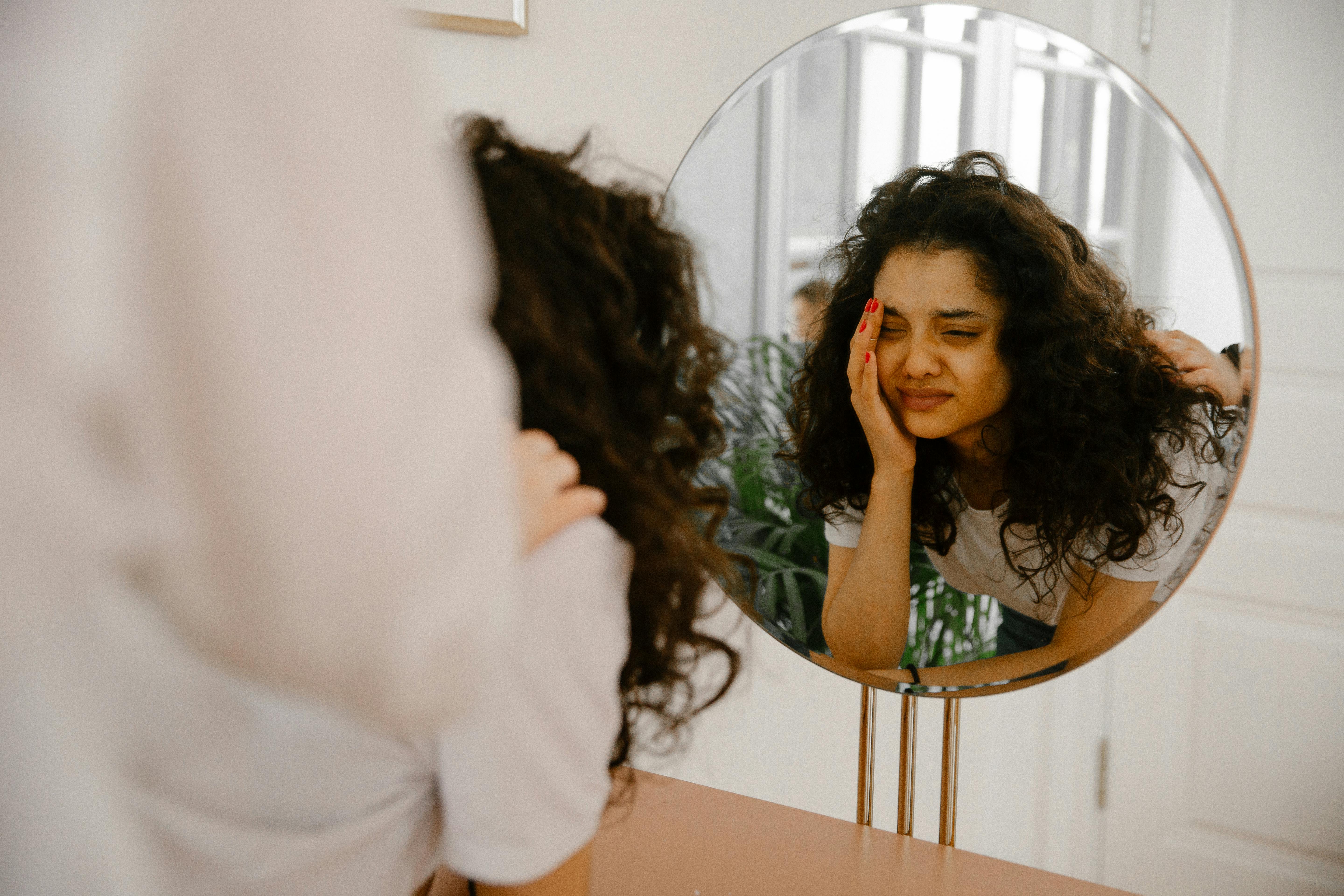 A woman looking thoughtfully into a mirror, resting her head on her hand, with a thoughtful expression.