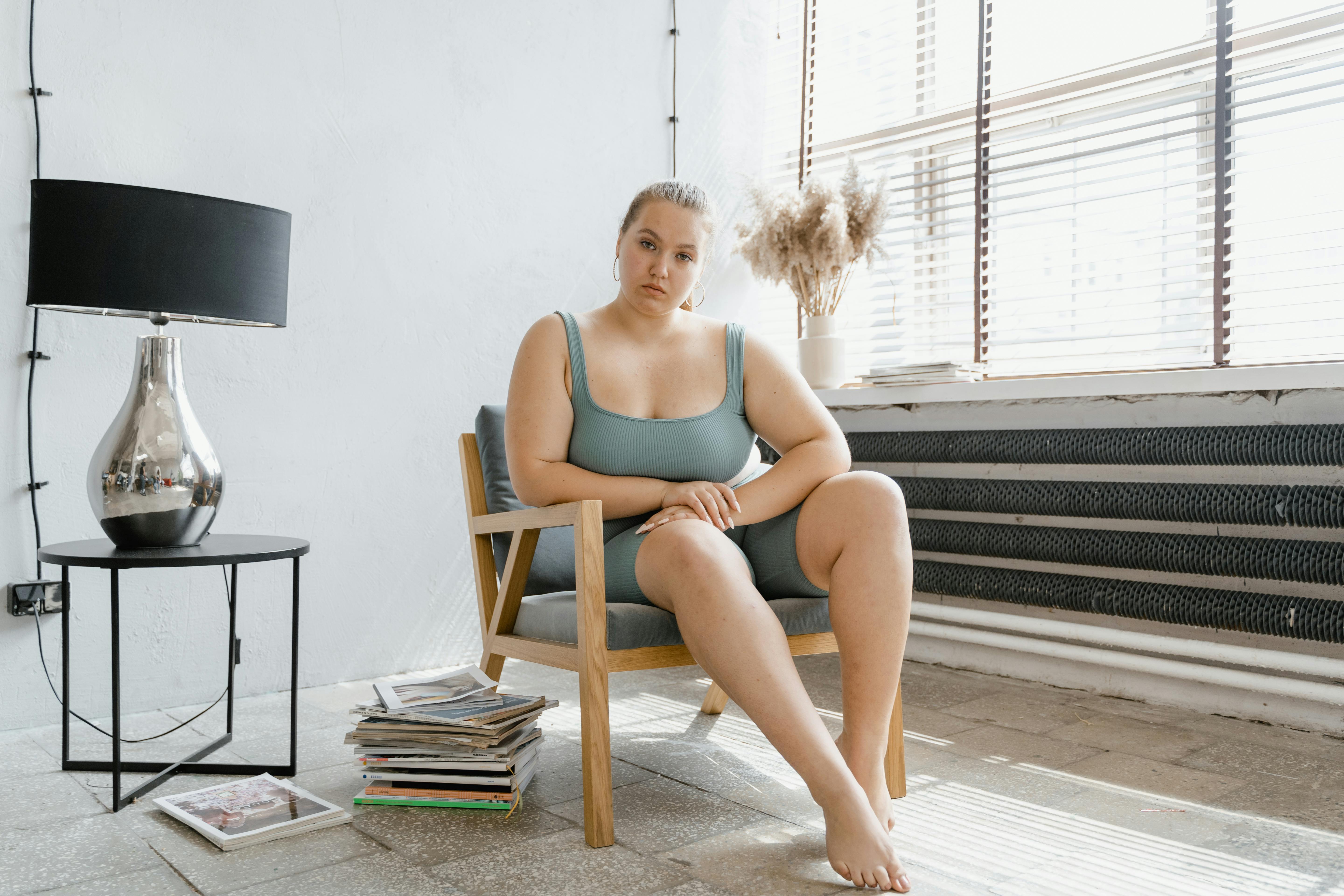 A woman in a teal outfit sitting on a wooden chair in a modern interior with a lamp and stacks of magazines.