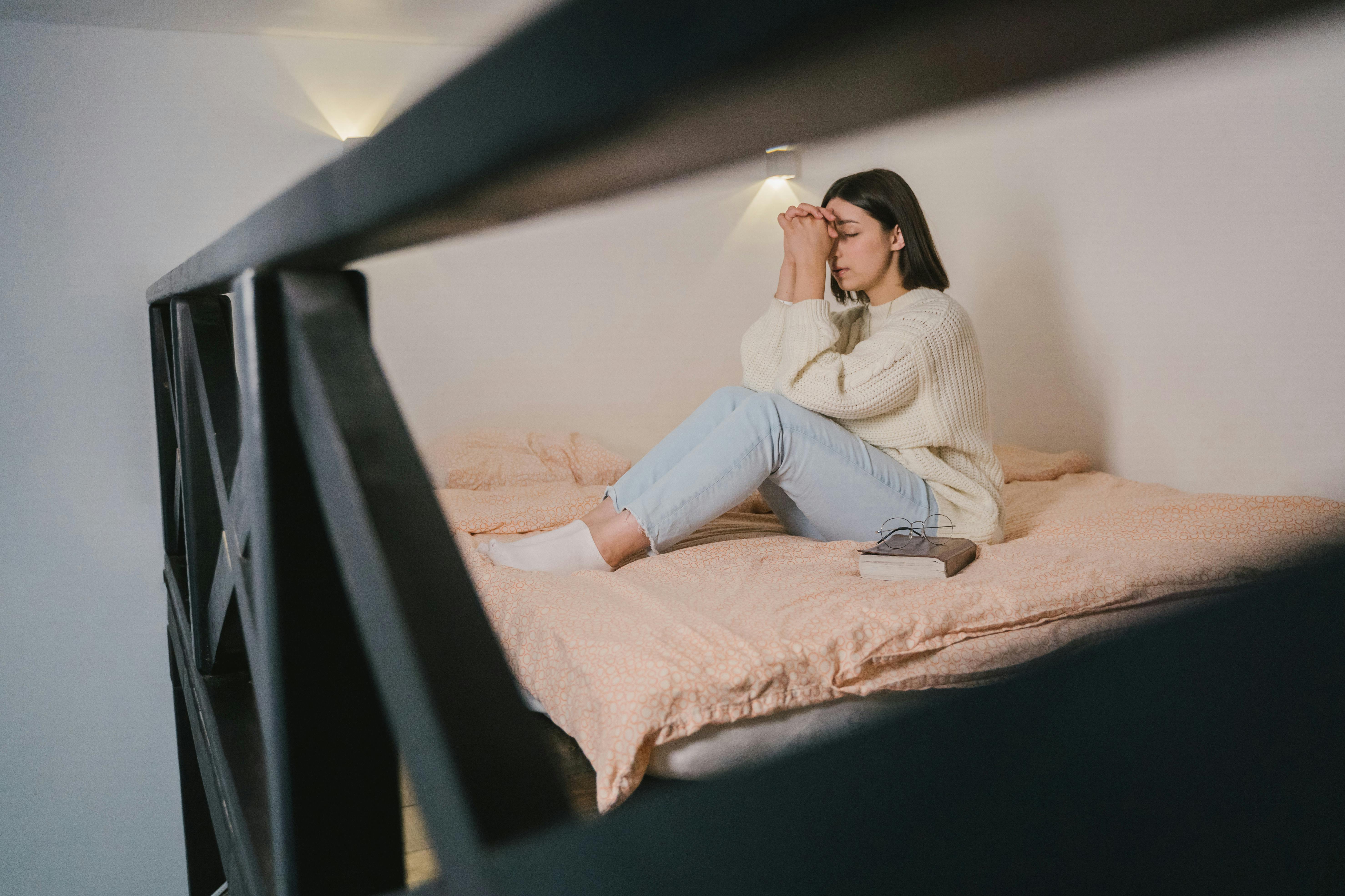 Woman sitting on a bed with her hands clasped in front of her face, looking contemplative.