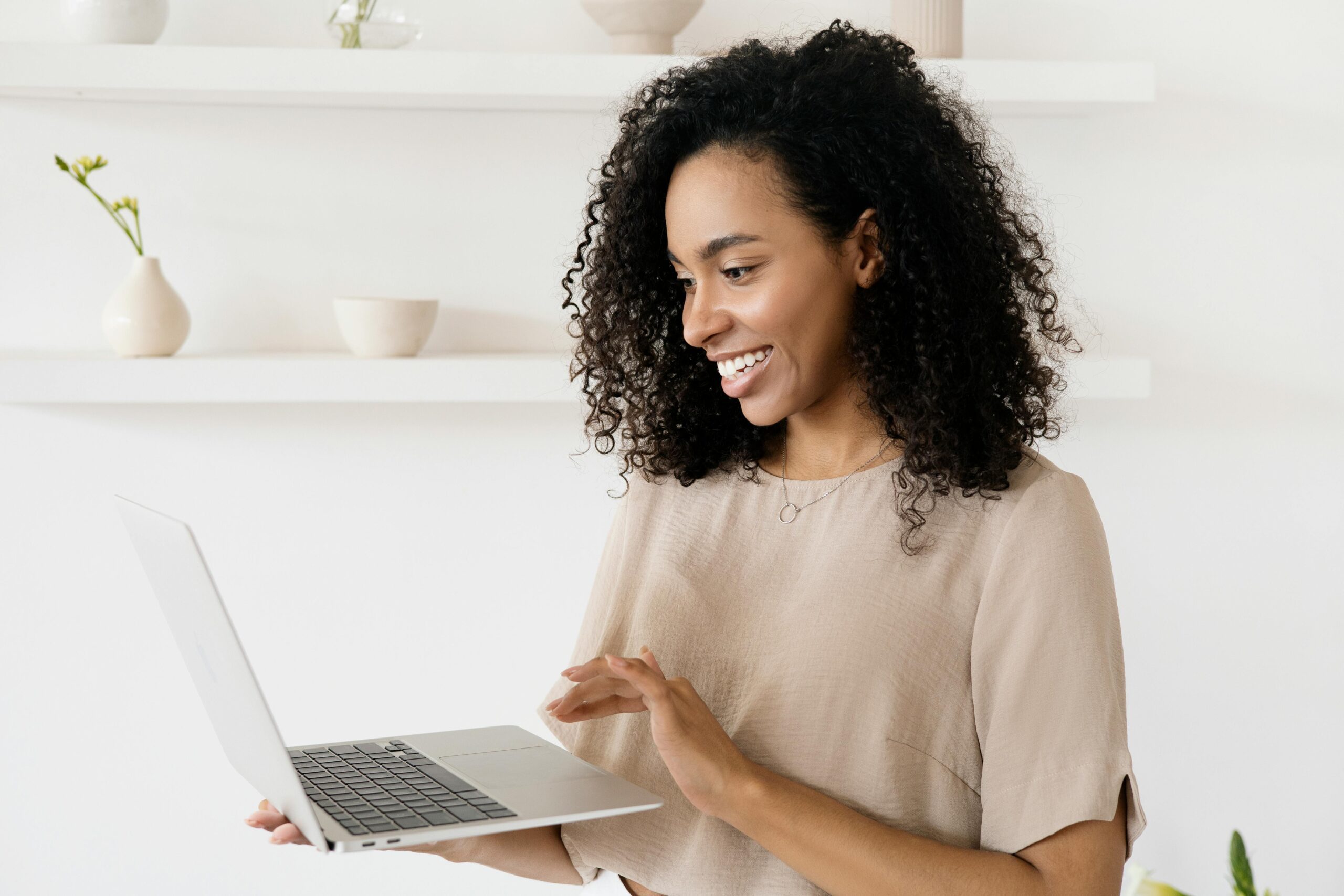 A woman with curly hair smiling while holding a laptop in a bright indoor setting.