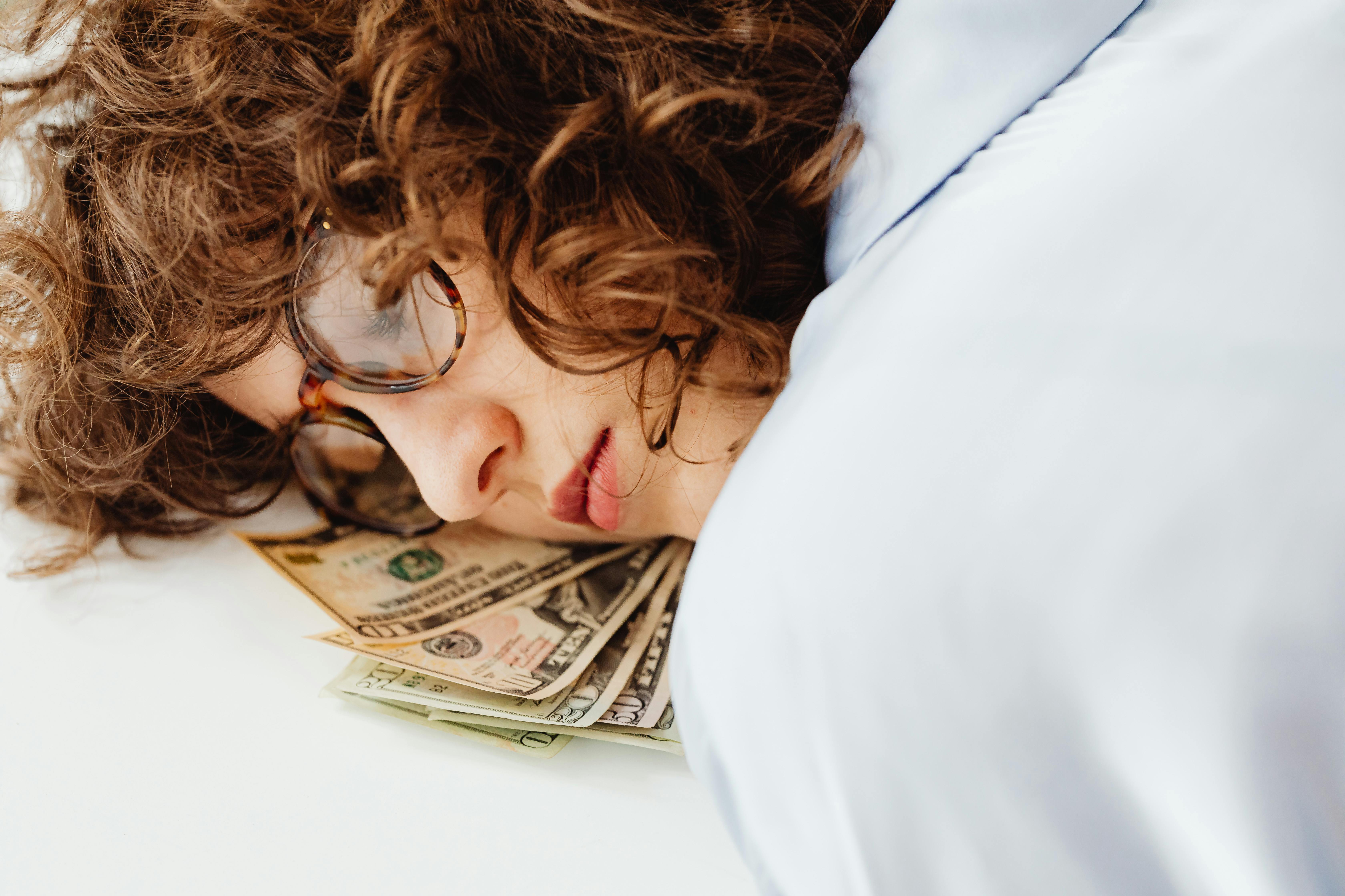 A woman laying her head on a pile of dollar bills, looking contemplative.