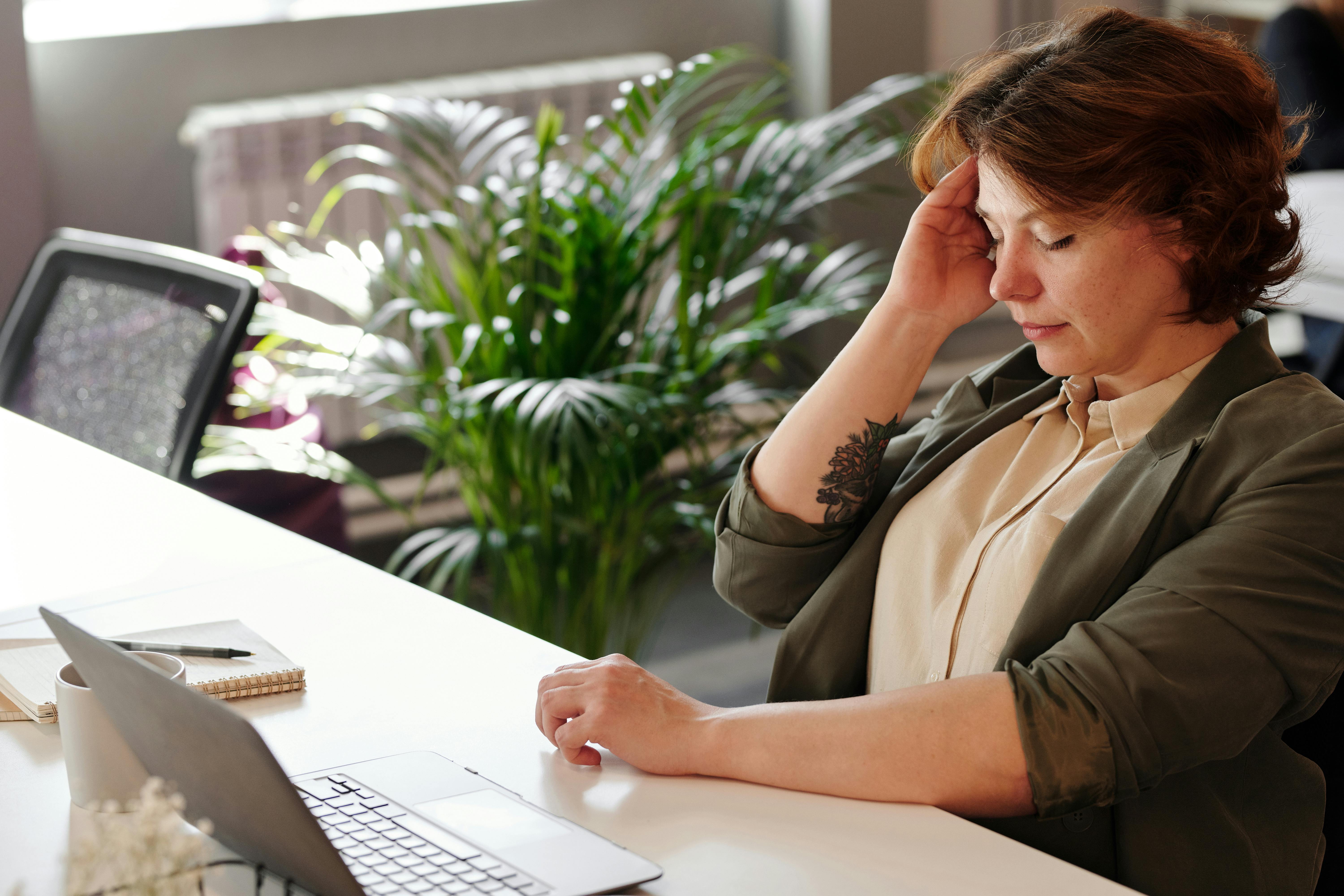 A woman with short hair sitting at a desk, looking stressed while holding her forehead in an office environment.