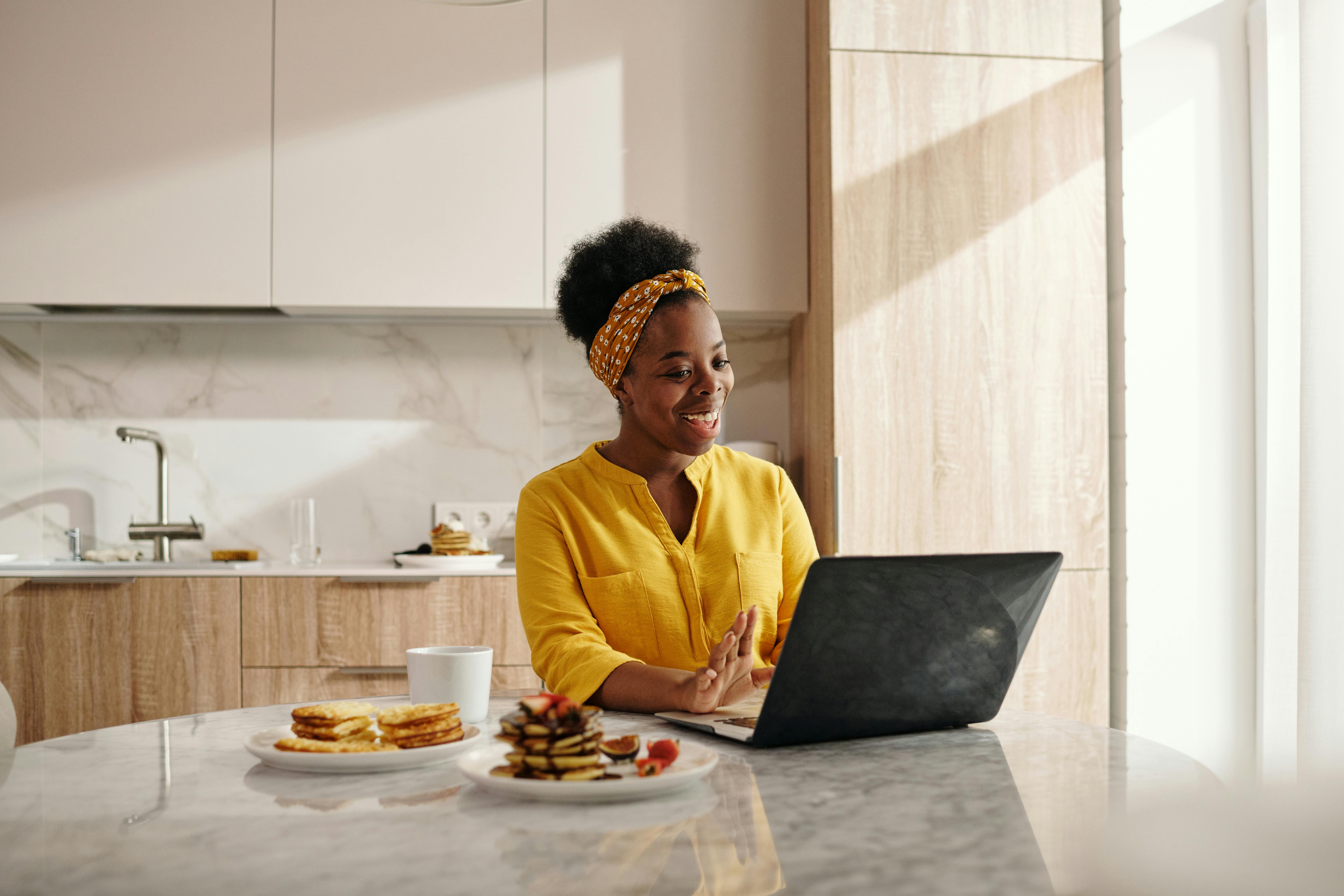 A woman in a yellow shirt works on a laptop at a kitchen table, smiling as she interacts on the screen.