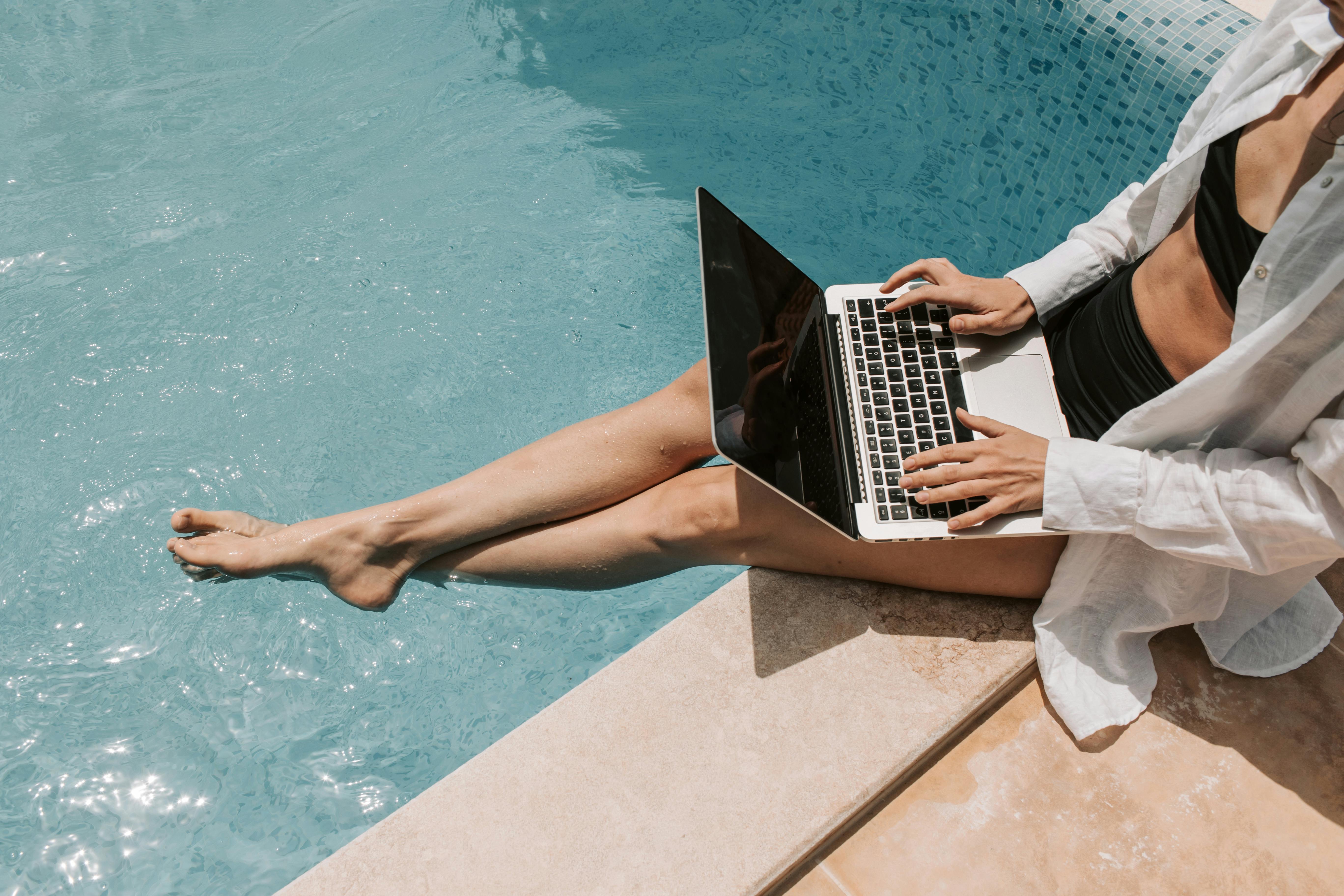 A woman sitting by the edge of a pool, using a laptop with her feet in the water.