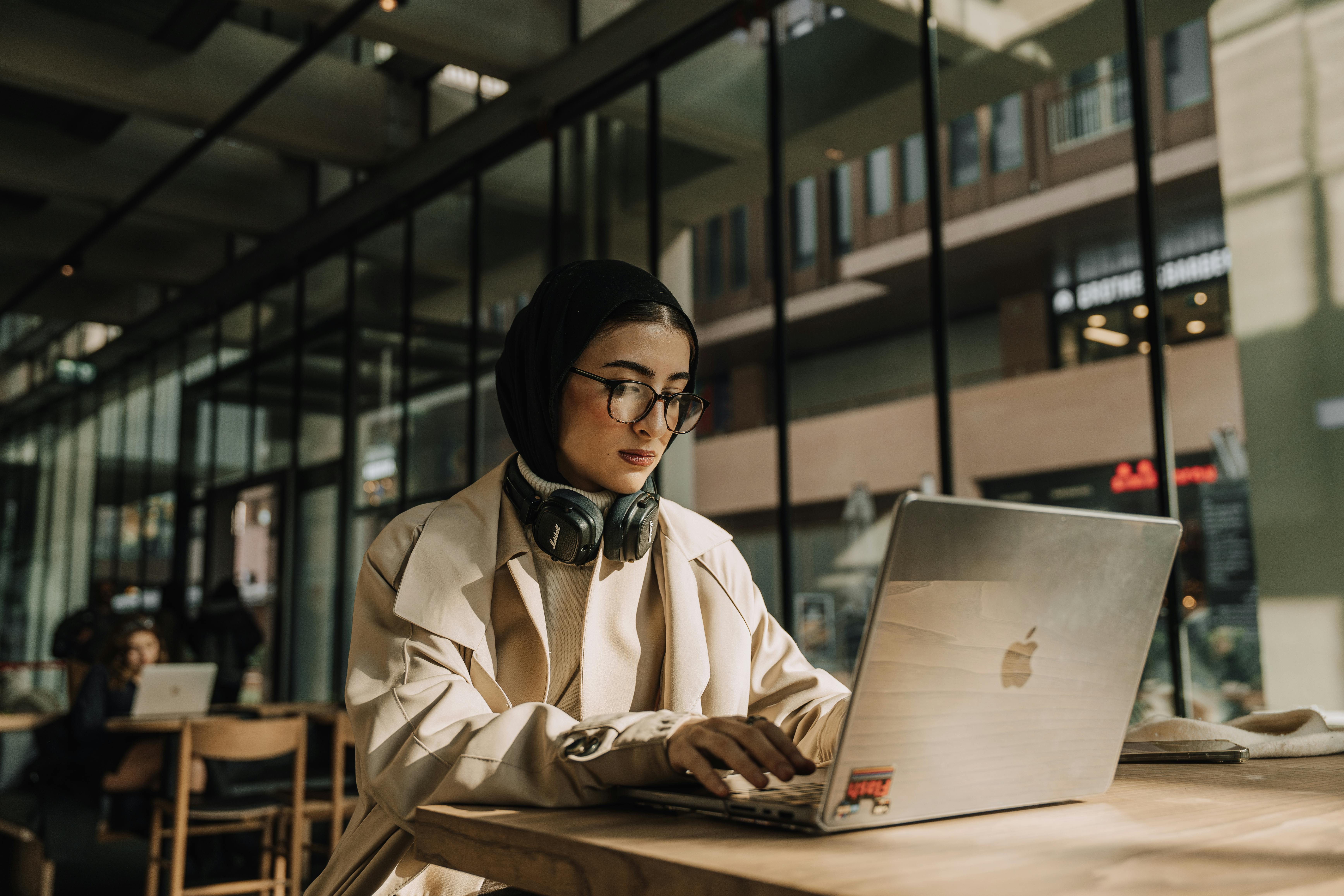 A woman wearing a hijab and glasses working on a laptop in a cafe