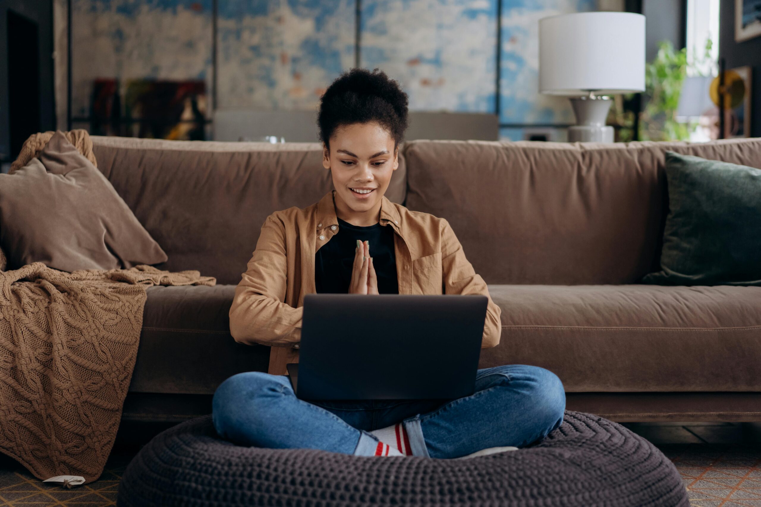 A woman sitting cross-legged on a large cushion working on a laptop in a cozy living room.